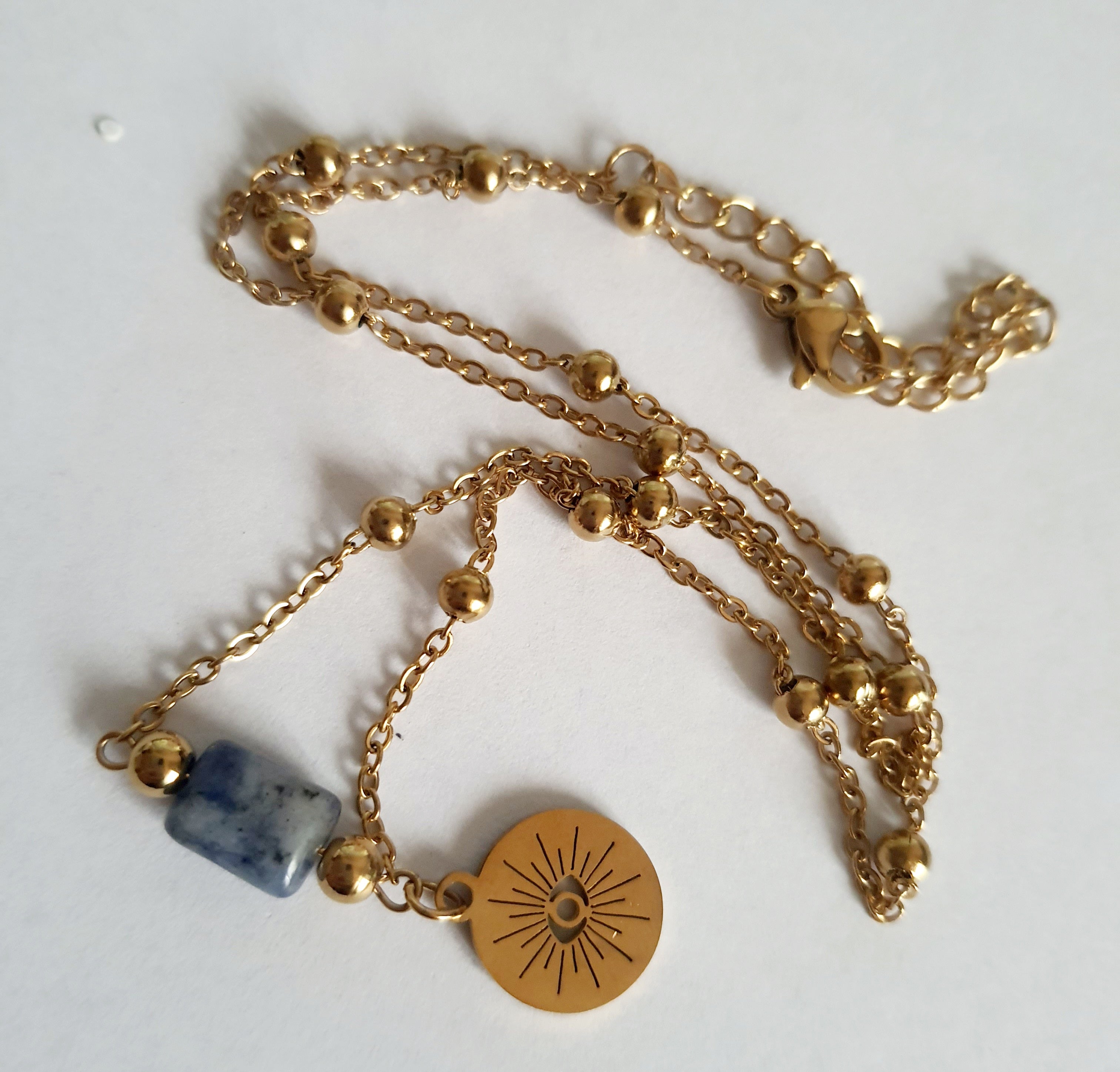 Złoty łańcuszek ze stali nierdzewnej o długości 40–45 cm z granatowym kamieniem i okrągłym talizmanem w kształcie słońca. Styl boho, minimalistyczny, duchowy. Rouge Bizu.