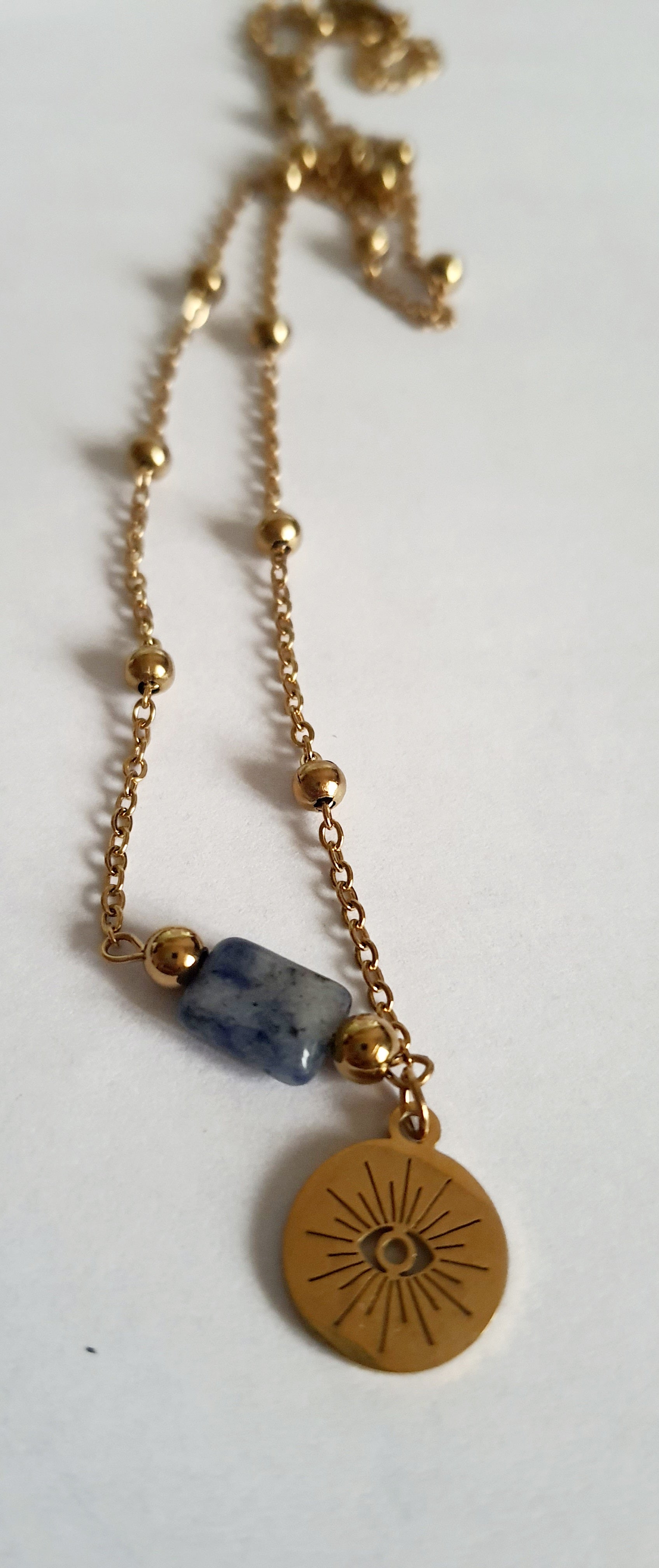 Złoty łańcuszek ze stali nierdzewnej o długości 40–45 cm z granatowym kamieniem i okrągłym talizmanem w kształcie słońca. Styl boho, minimalistyczny, duchowy. Rouge Bizu.