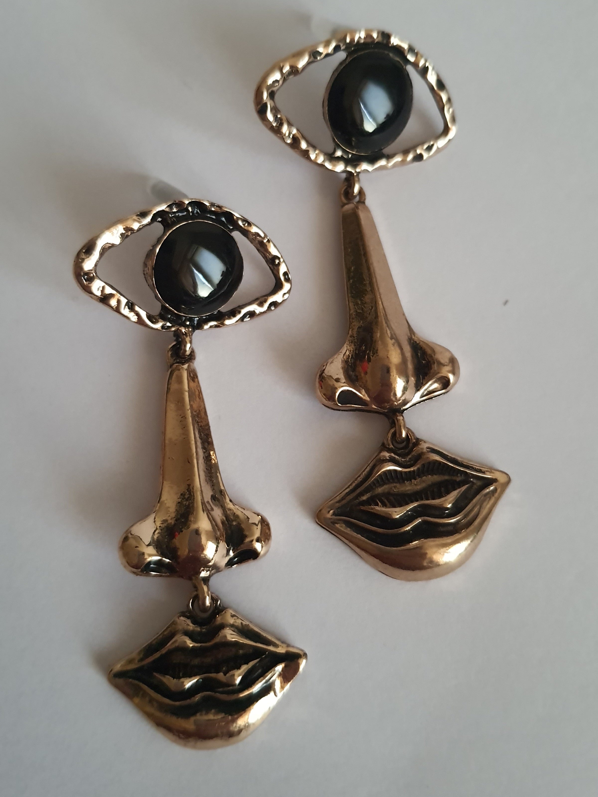 Wiszące kolczyki 7 cm ze stali nierdzewnej w kolorze złotym w kształcie oka, nosa i ust, z czarnym kamieniem w centrum oka. Rouge Bizu, stal nierdzewna, stal chirurgiczna.