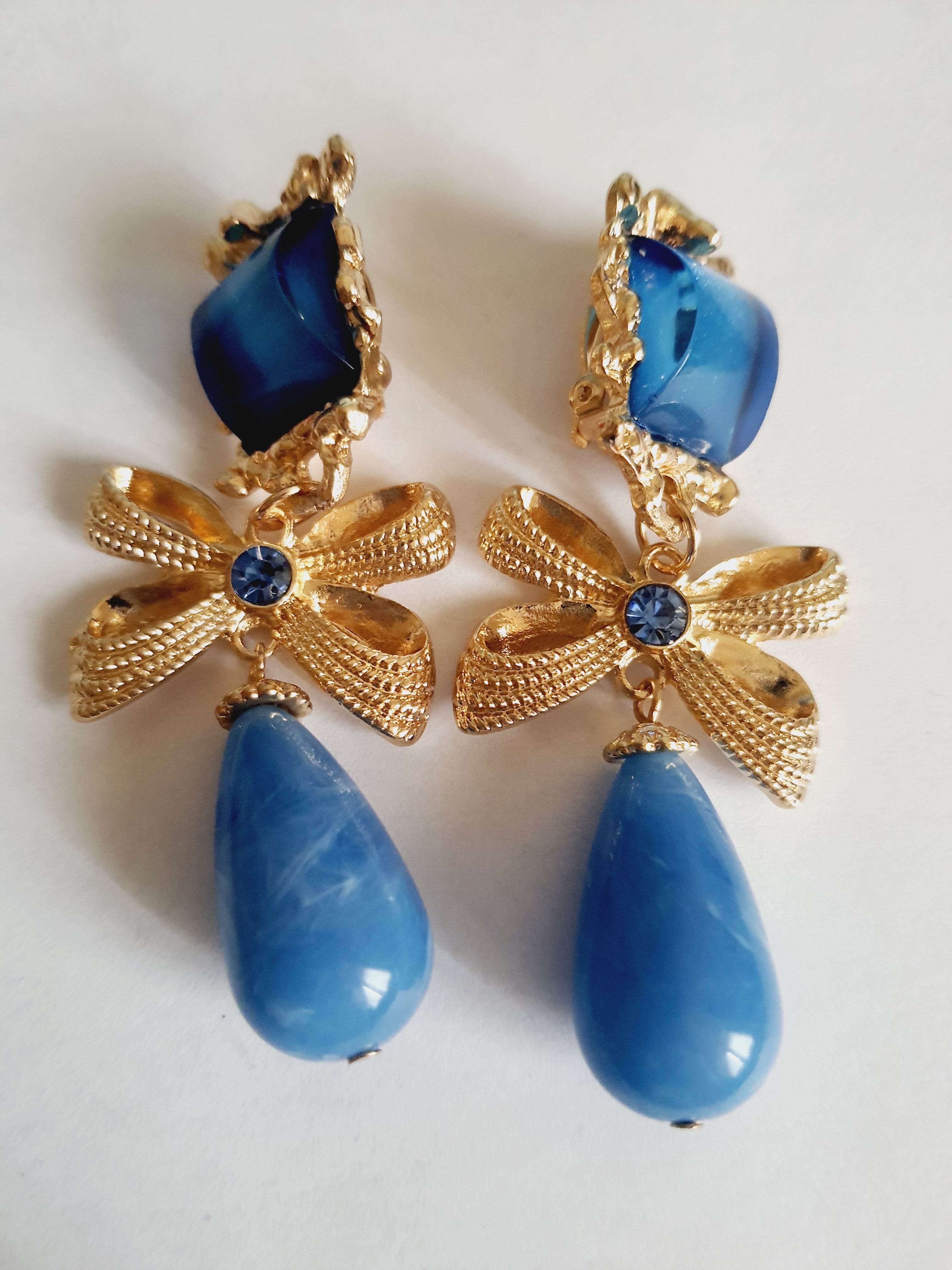 Złote klipsy ze stali nierdzewnej z niebieskimi kryształami. Dekoracyjne złote kokardy z błękitnym kamieniem i dużą kroplą w odcieniu niebieskiego. Styl glamour, romantyczna elegancja. Długość 7 cm. Rouge Bizu.