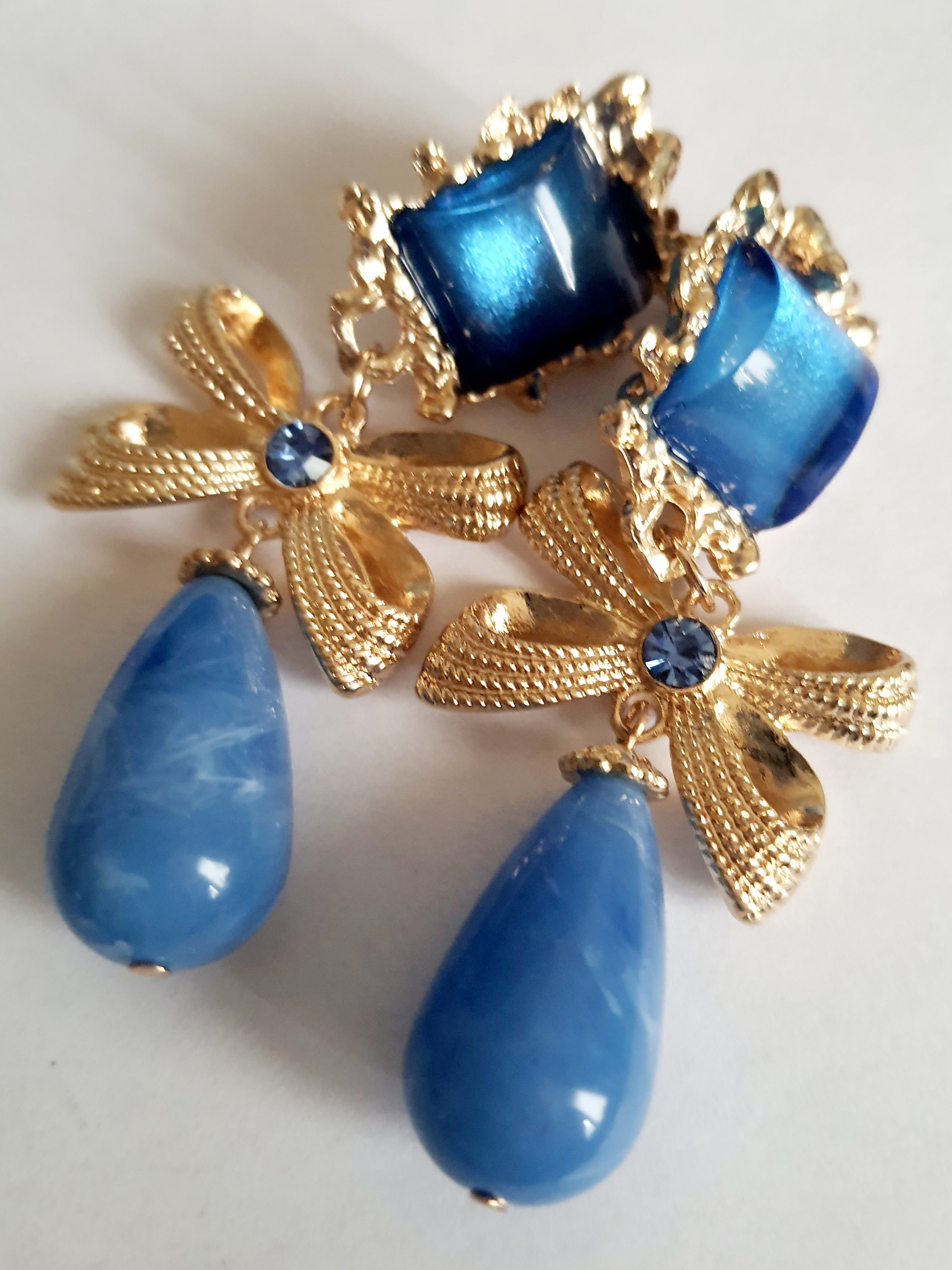 Złote klipsy ze stali nierdzewnej z niebieskimi kryształami. Dekoracyjne złote kokardy z błękitnym kamieniem i dużą kroplą w odcieniu niebieskiego. Styl glamour, romantyczna elegancja. Długość 7 cm. Rouge Bizu.