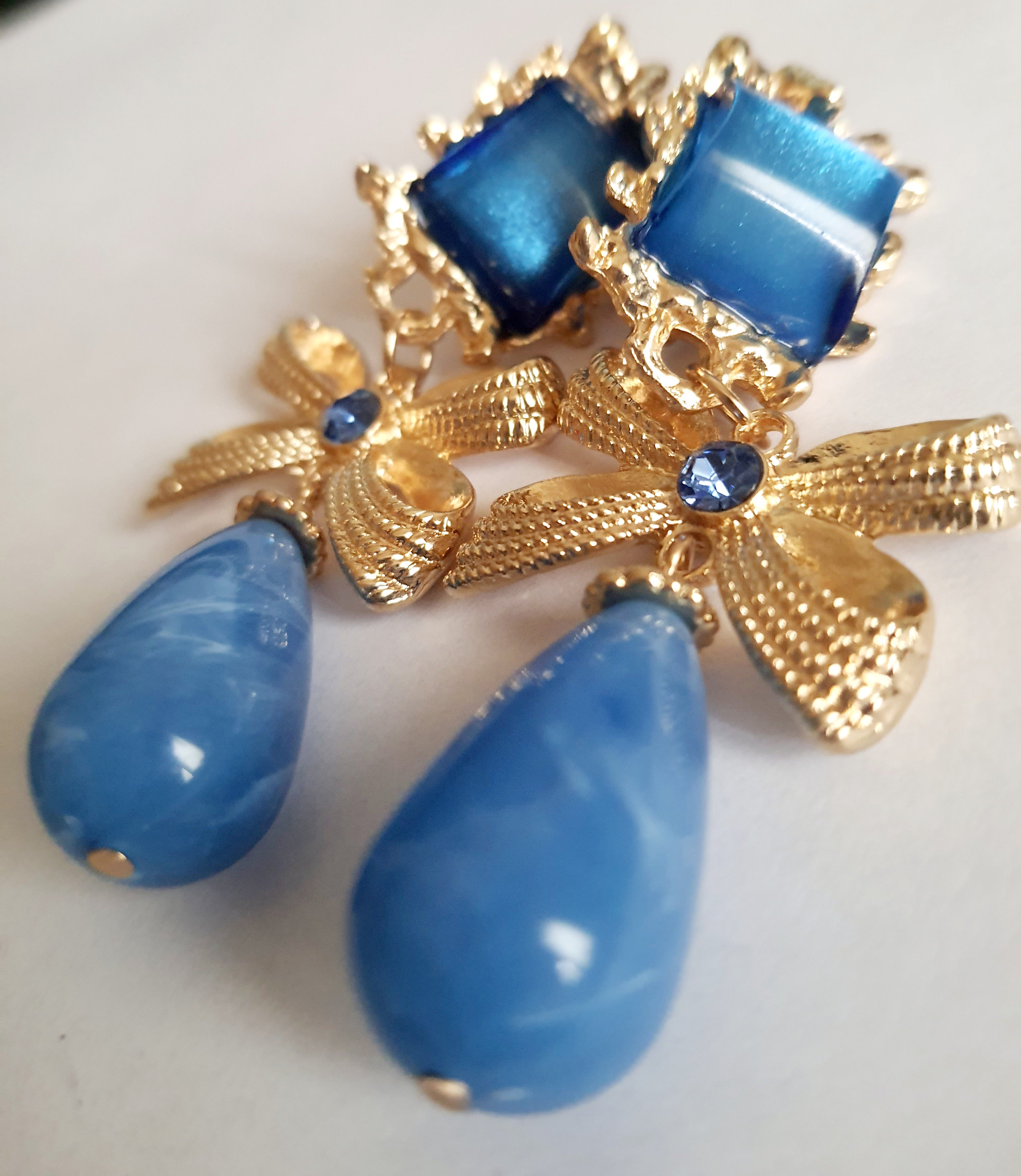 Złote klipsy ze stali nierdzewnej z niebieskimi kryształami. Dekoracyjne złote kokardy z błękitnym kamieniem i dużą kroplą w odcieniu niebieskiego. Styl glamour, romantyczna elegancja. Długość 7 cm. Rouge Bizu.