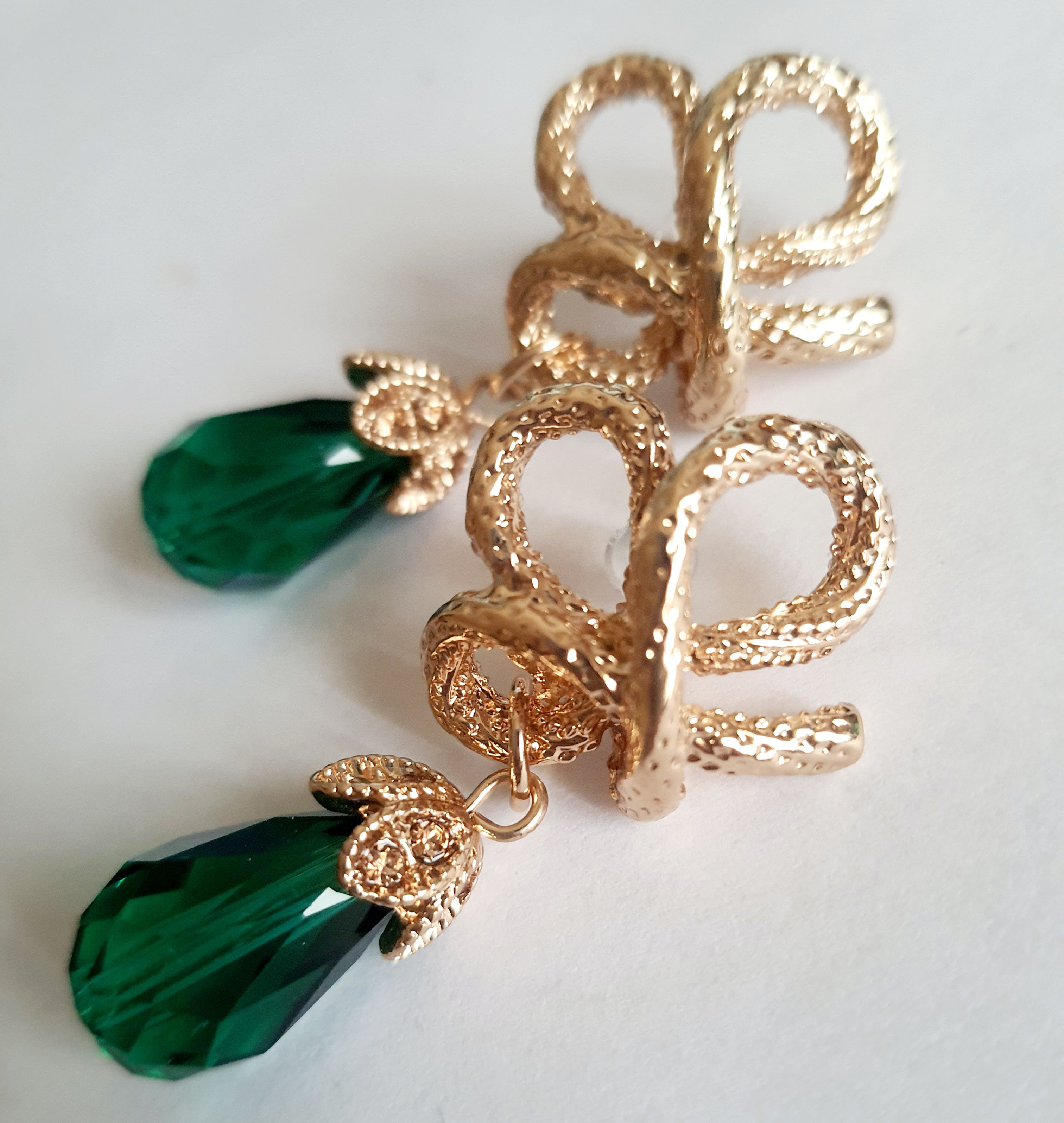 Złote kolczyki ze stali nierdzewnej w stylu glamour, ozdobione rzeźbioną kokardą i zielonym kryształem w kształcie kropli. Elegancja i szmaragdowy blask. Długość 6 cm. Rouge Bizu.