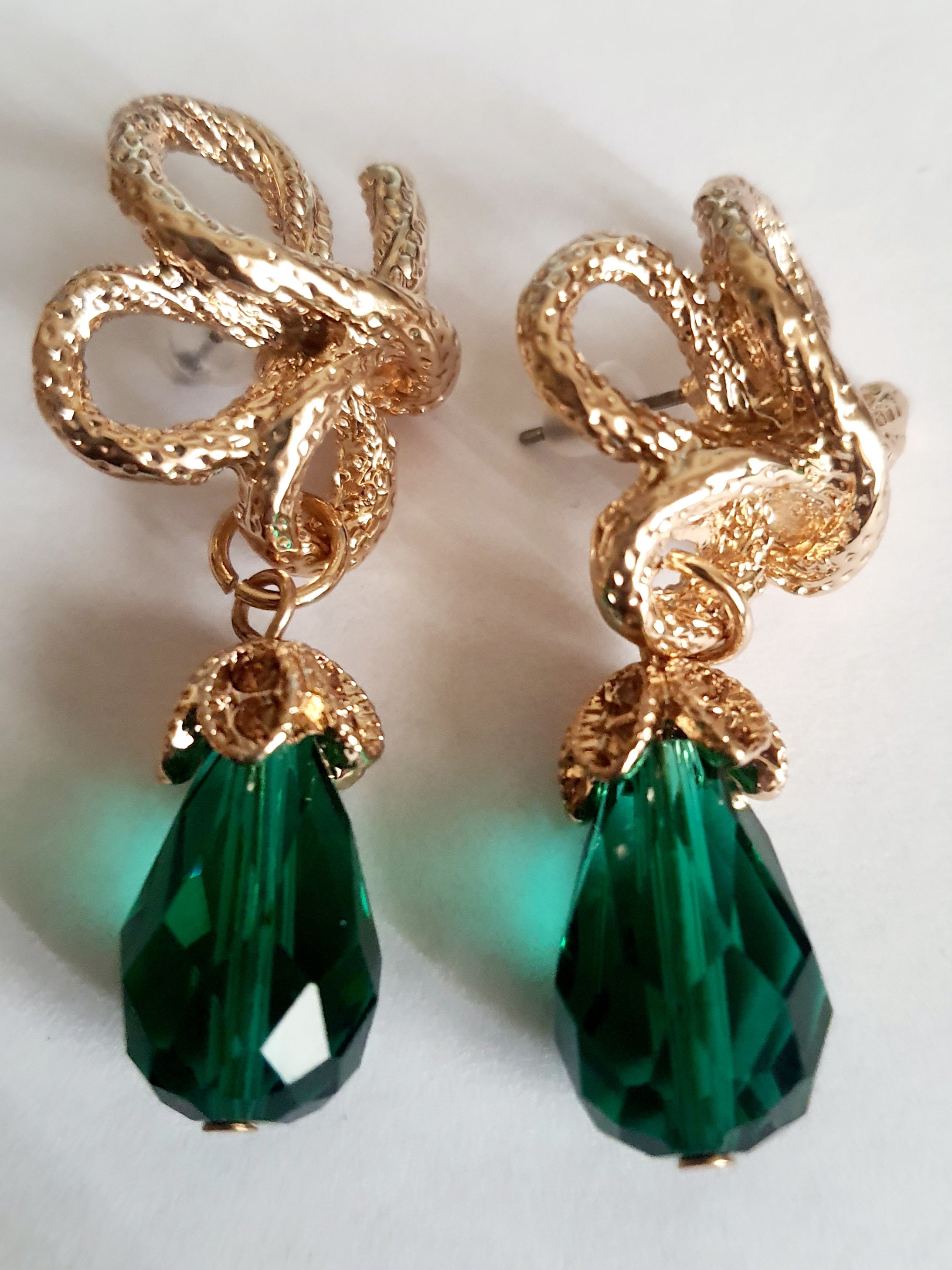 Złote kolczyki ze stali nierdzewnej w stylu glamour, ozdobione rzeźbioną kokardą i zielonym kryształem w kształcie kropli. Elegancja i szmaragdowy blask. Długość 6 cm. Rouge Bizu.