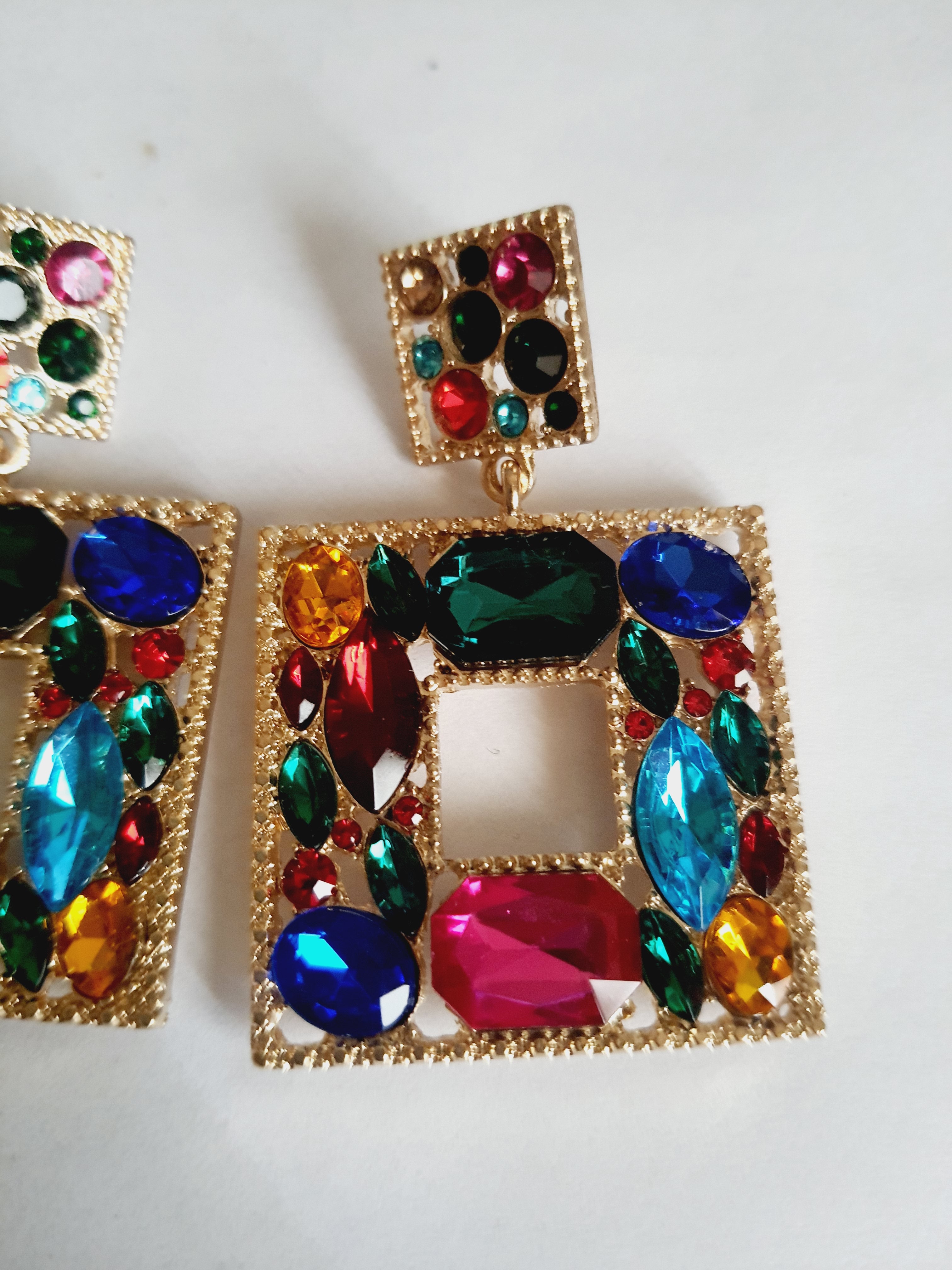 Złote kolczyki ze stali nierdzewnej w kształcie kwadratu, bogato zdobione wielobarwnymi kryształami w odcieniach granatu, zieleni, różu, czerwieni, turkusu i złota. Styl glamour, długość 6 cm. Rouge Bizu.