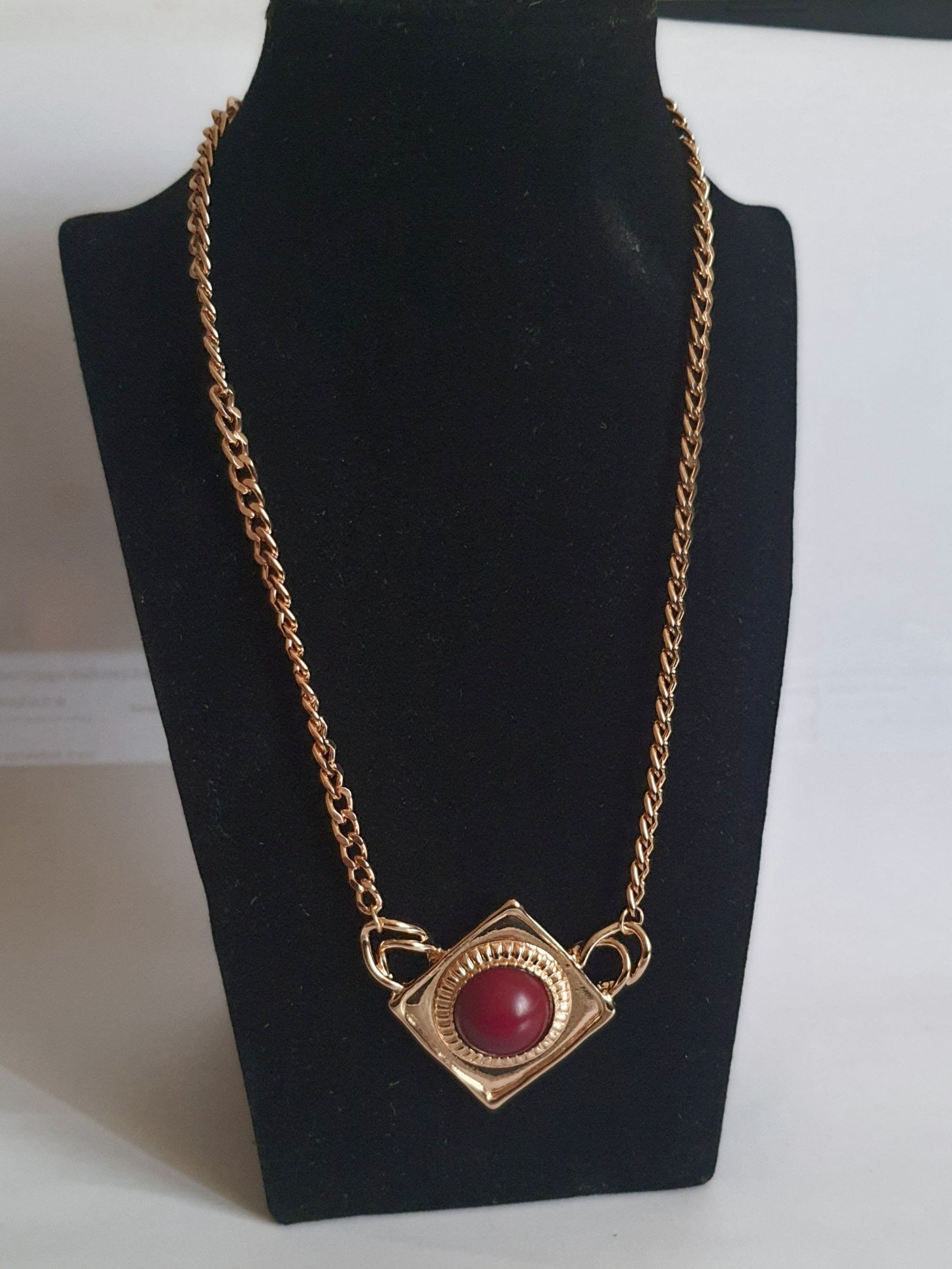 Naszyjnik ze stali nierdzewnej w kolorze złota, z geometryczną zawieszką w kształcie rombu i czerwonym kamieniem pośrodku. Styl elegancki, kobiecy, długość regulowana 44,5–54,5 cm. Rouge Bizu.