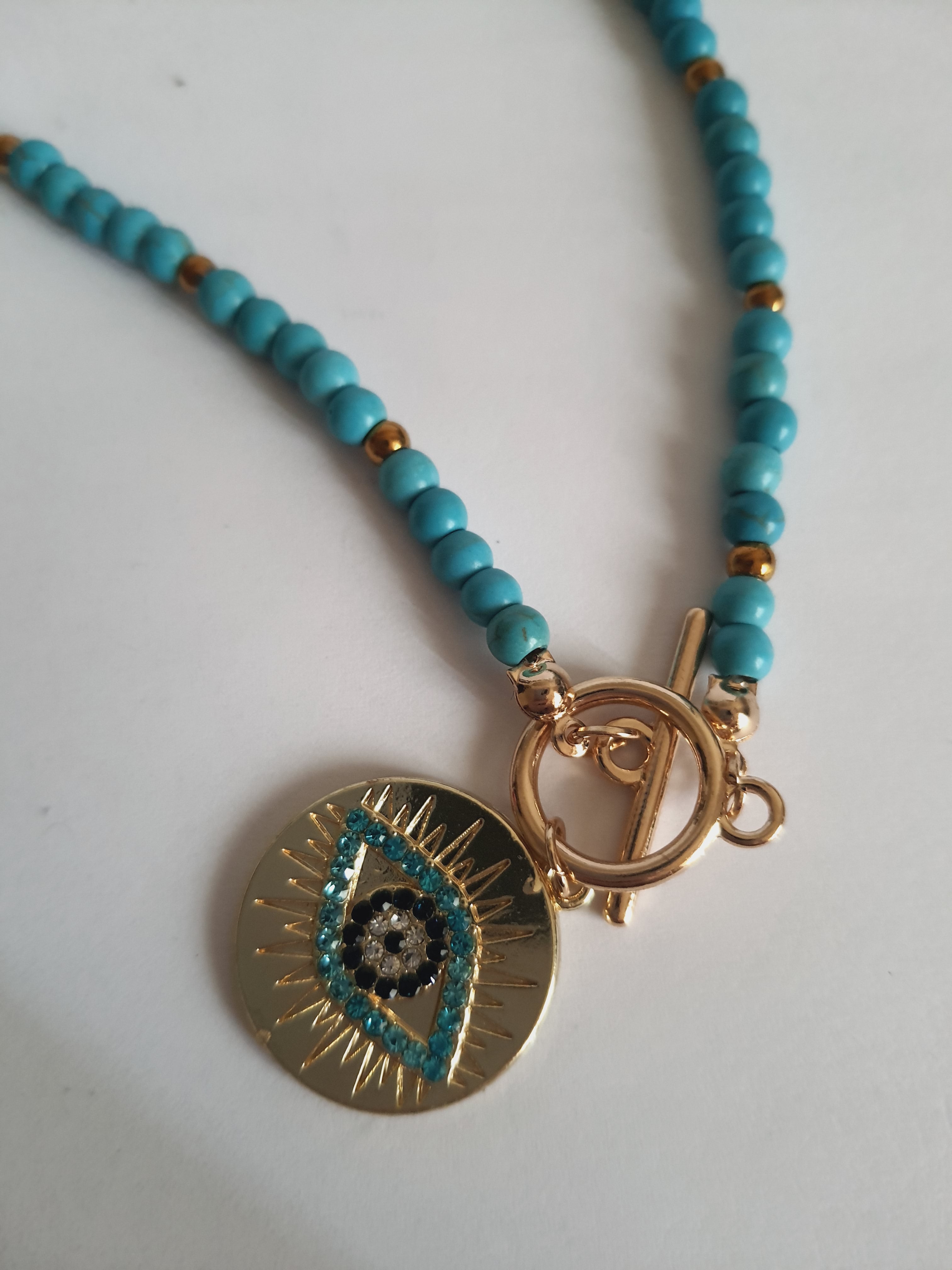 Naszyjnik choker z turkusowych koralików i złotych przekładek, z okrągłą złotą zawieszką z motywem oka w odcieniach turkusu i czerni. Długość 40 cm. Stal nierdzewna. Rouge Bizu.