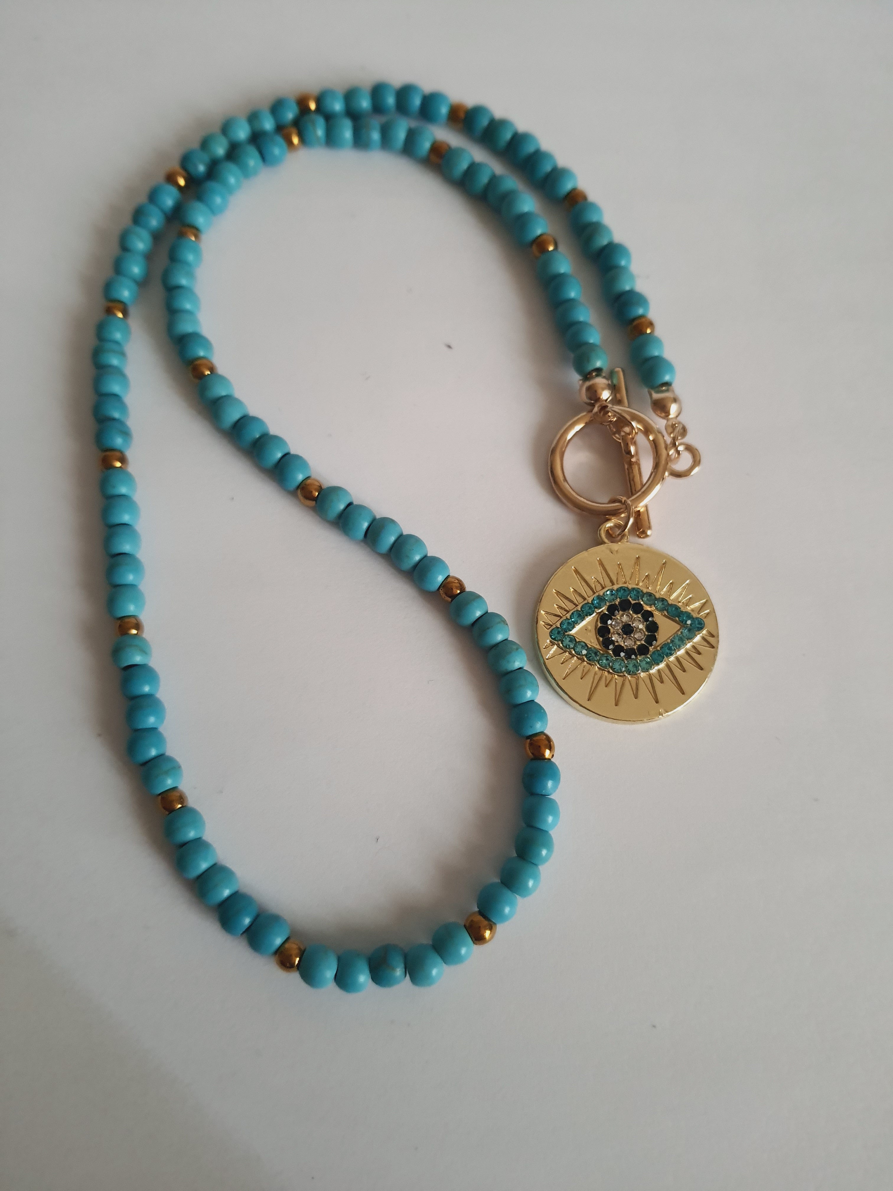 Naszyjnik choker z turkusowych koralików i złotych przekładek, z okrągłą złotą zawieszką z motywem oka w odcieniach turkusu i czerni. Długość 40 cm. Stal nierdzewna. Rouge Bizu.