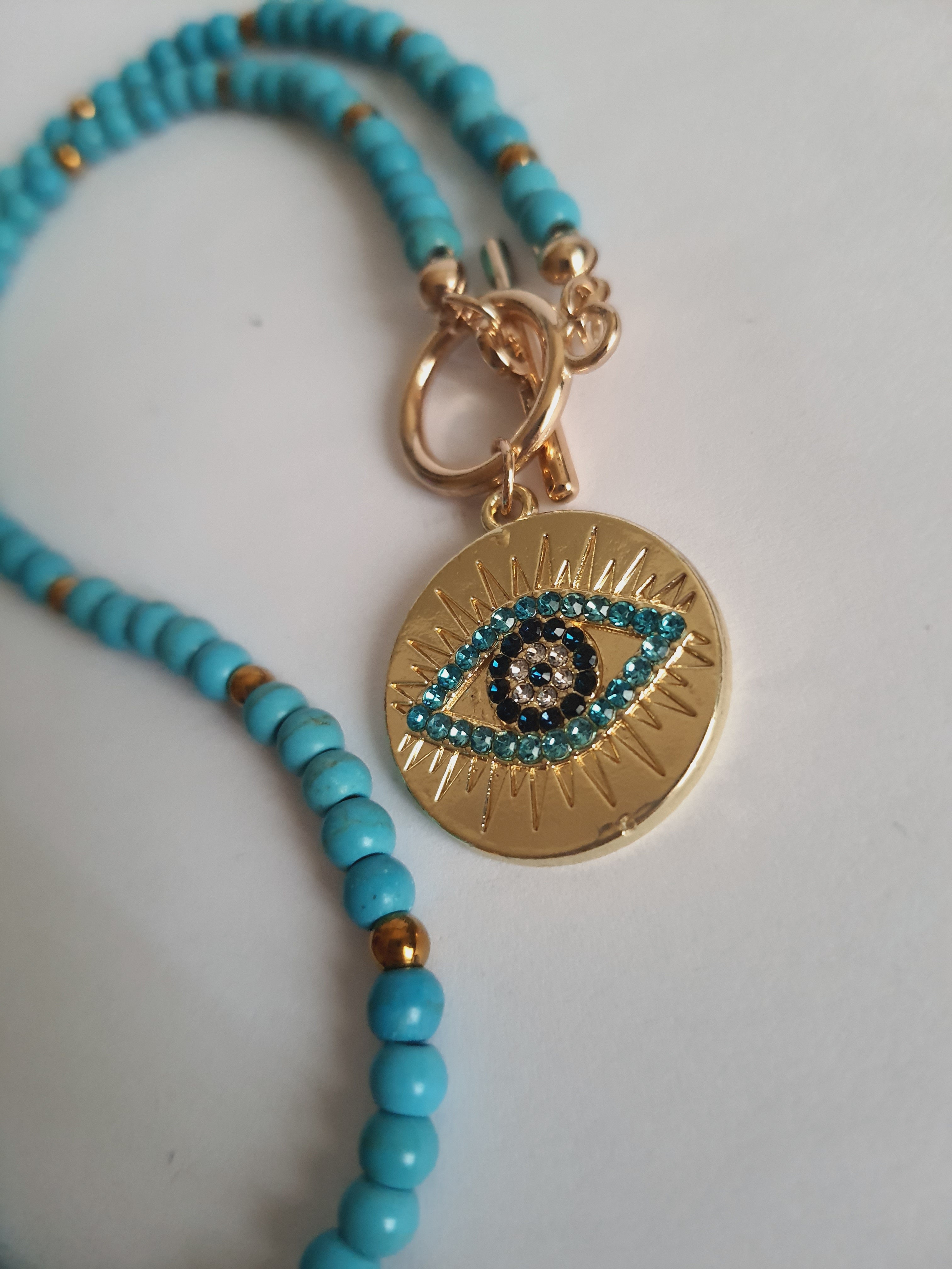 Naszyjnik choker z turkusowych koralików i złotych przekładek, z okrągłą złotą zawieszką z motywem oka w odcieniach turkusu i czerni. Długość 40 cm. Stal nierdzewna. Rouge Bizu.