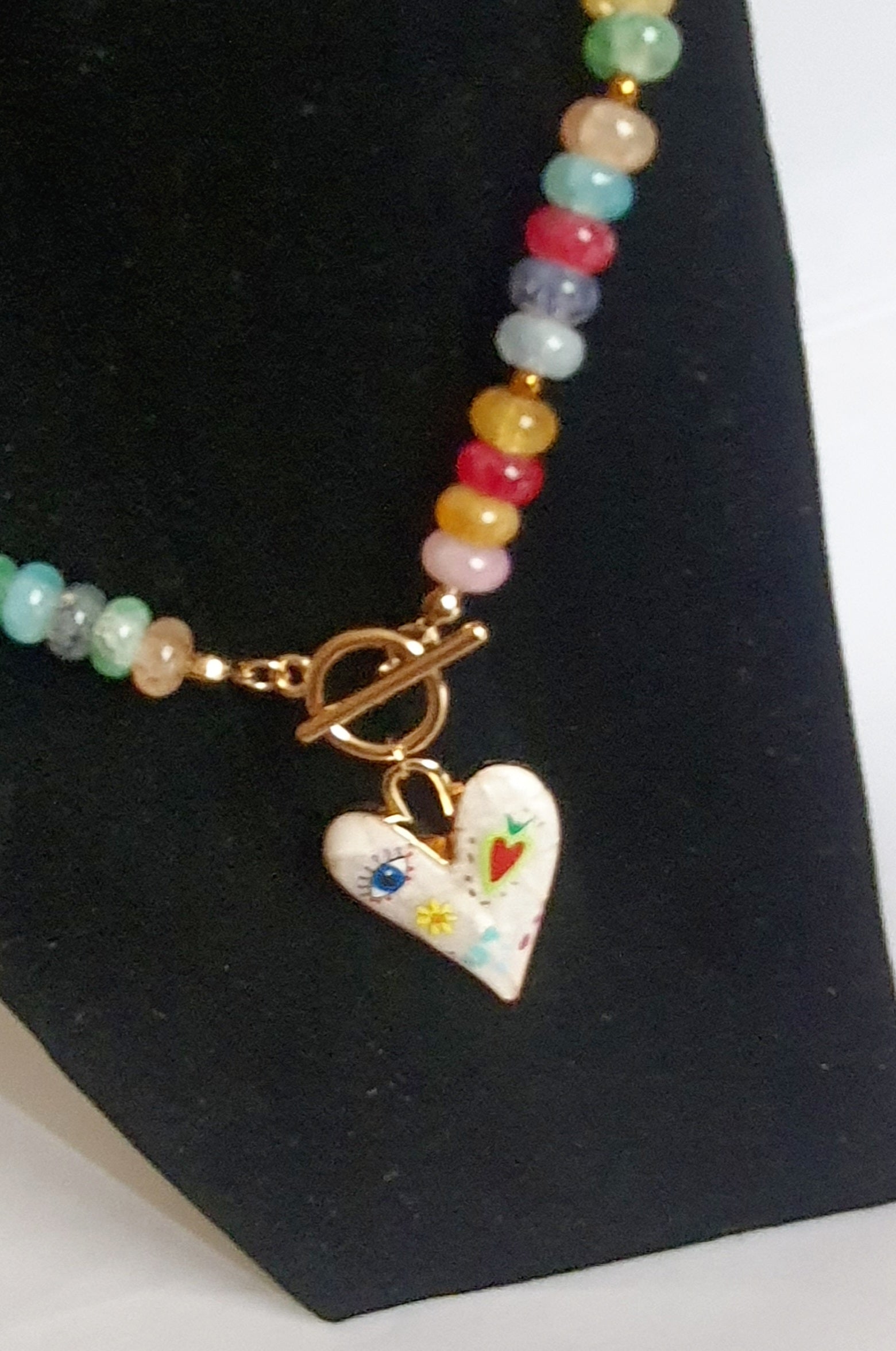 Naszyjnik z kolorowych koralików w stylu boho, z ozdobną zawieszką w kształcie białego serca zdobionego ręcznie malowanymi wzorami: oko, kwiat, serce i kolorowe symbole. Długość 45 cm. Stal nierdzewna. Rouge Bizu.