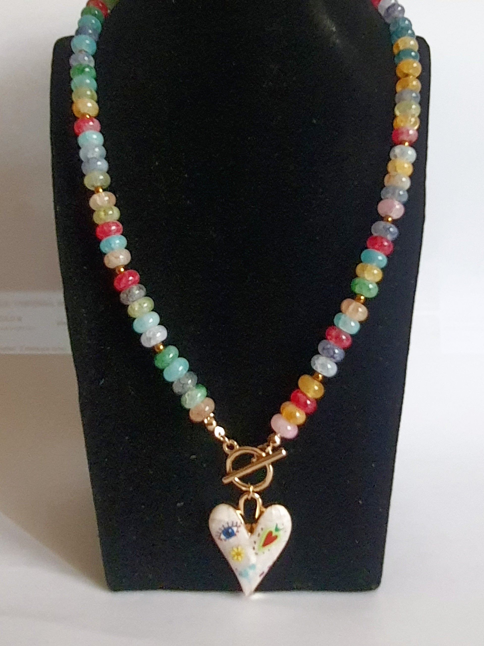 Naszyjnik z kolorowych koralików w stylu boho, z ozdobną zawieszką w kształcie białego serca zdobionego ręcznie malowanymi wzorami: oko, kwiat, serce i kolorowe symbole. Długość 45 cm. Stal nierdzewna. Rouge Bizu.