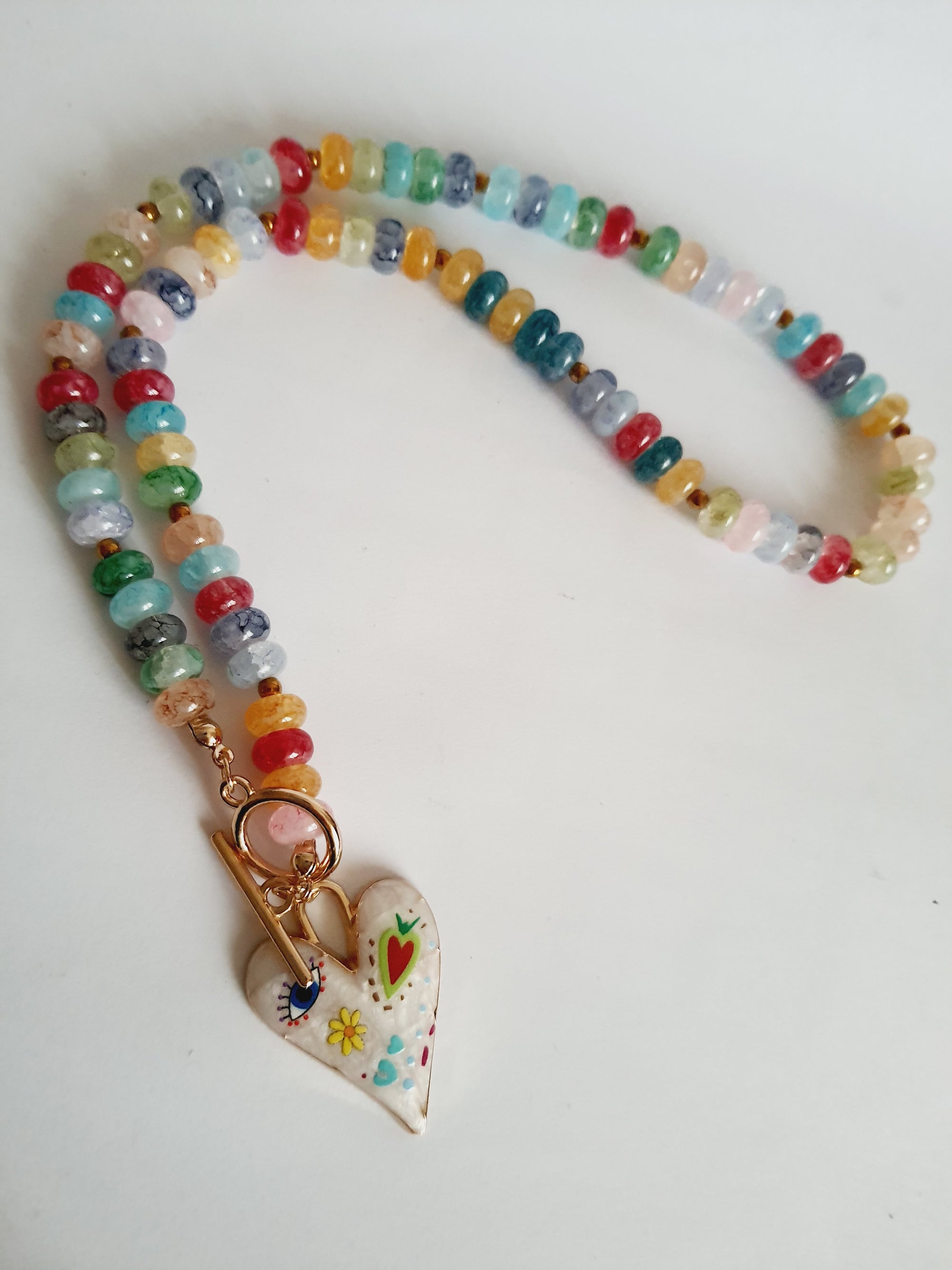 Naszyjnik z kolorowych koralików w stylu boho, z ozdobną zawieszką w kształcie białego serca zdobionego ręcznie malowanymi wzorami: oko, kwiat, serce i kolorowe symbole. Długość 45 cm. Stal nierdzewna. Rouge Bizu.