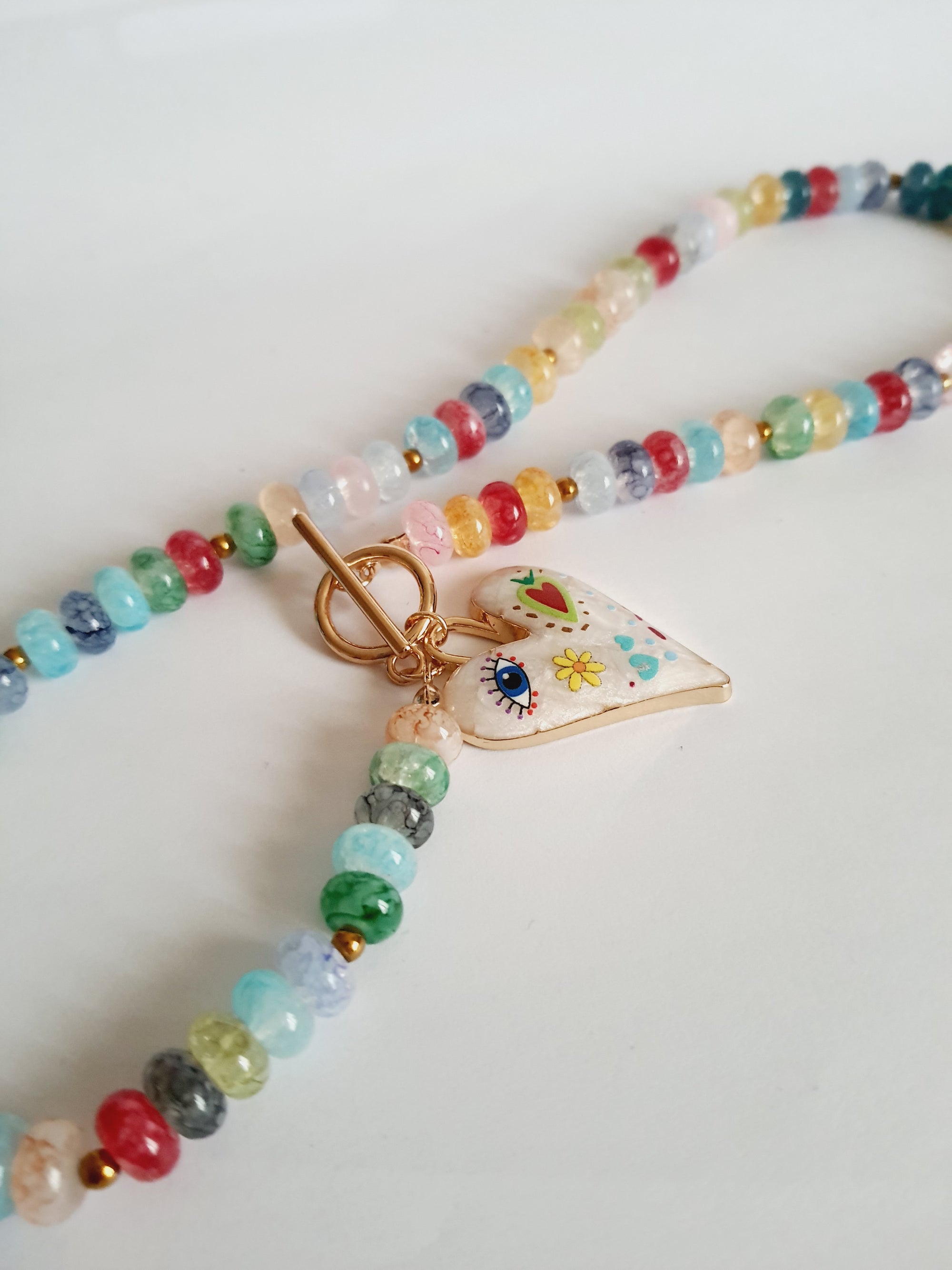 Naszyjnik z kolorowych koralików w stylu boho, z ozdobną zawieszką w kształcie białego serca zdobionego ręcznie malowanymi wzorami: oko, kwiat, serce i kolorowe symbole. Długość 45 cm. Stal nierdzewna. Rouge Bizu.