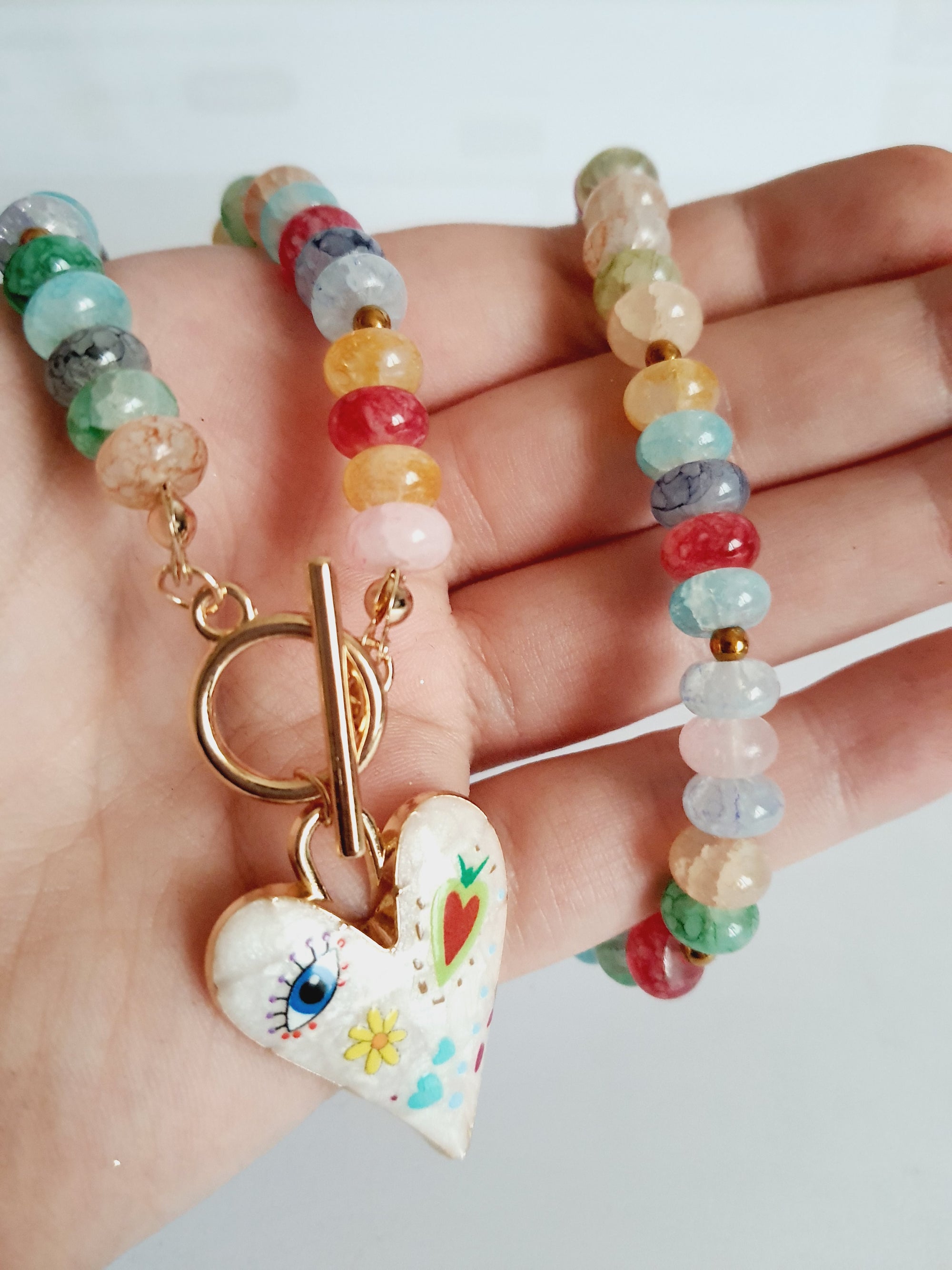 Naszyjnik z kolorowych koralików w stylu boho, z ozdobną zawieszką w kształcie białego serca zdobionego ręcznie malowanymi wzorami: oko, kwiat, serce i kolorowe symbole. Długość 45 cm. Stal nierdzewna. Rouge Bizu.