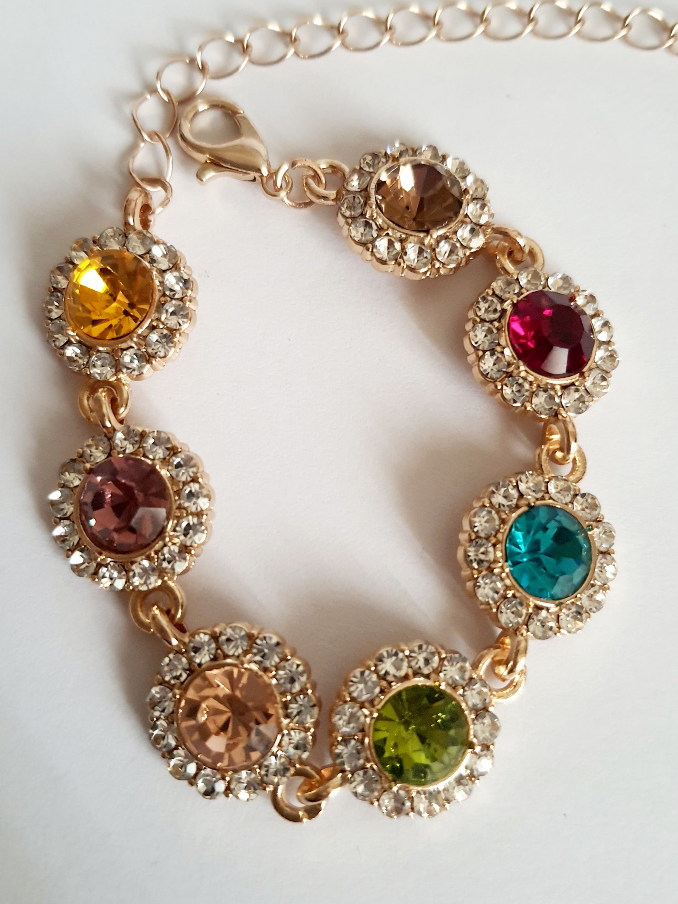 Bransoletka ze stali nierdzewnej w kolorze złota, z okrągłymi kolorowymi kryształami w odcieniach czerwieni, turkusu, zieleni, złota i bursztynu, otoczonymi małymi cyrkoniami. Długość regulowana 25 cm. Rouge Bizu.