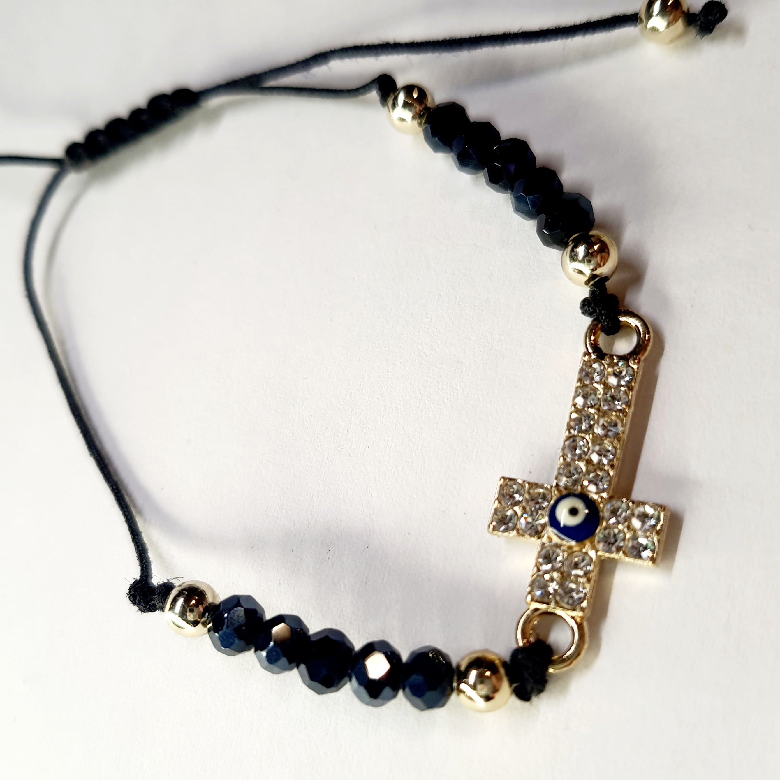 Bransoletka z krzyżem wysadzanym cyrkoniami i okiem proroka w złotej oprawie, na czarnym sznurku z ciemnymi koralikami. Ochronna i elegancka biżuteria w stylu boho. Wodoodporne, hipoalergiczne, jakość premium. Rouge Bizu.