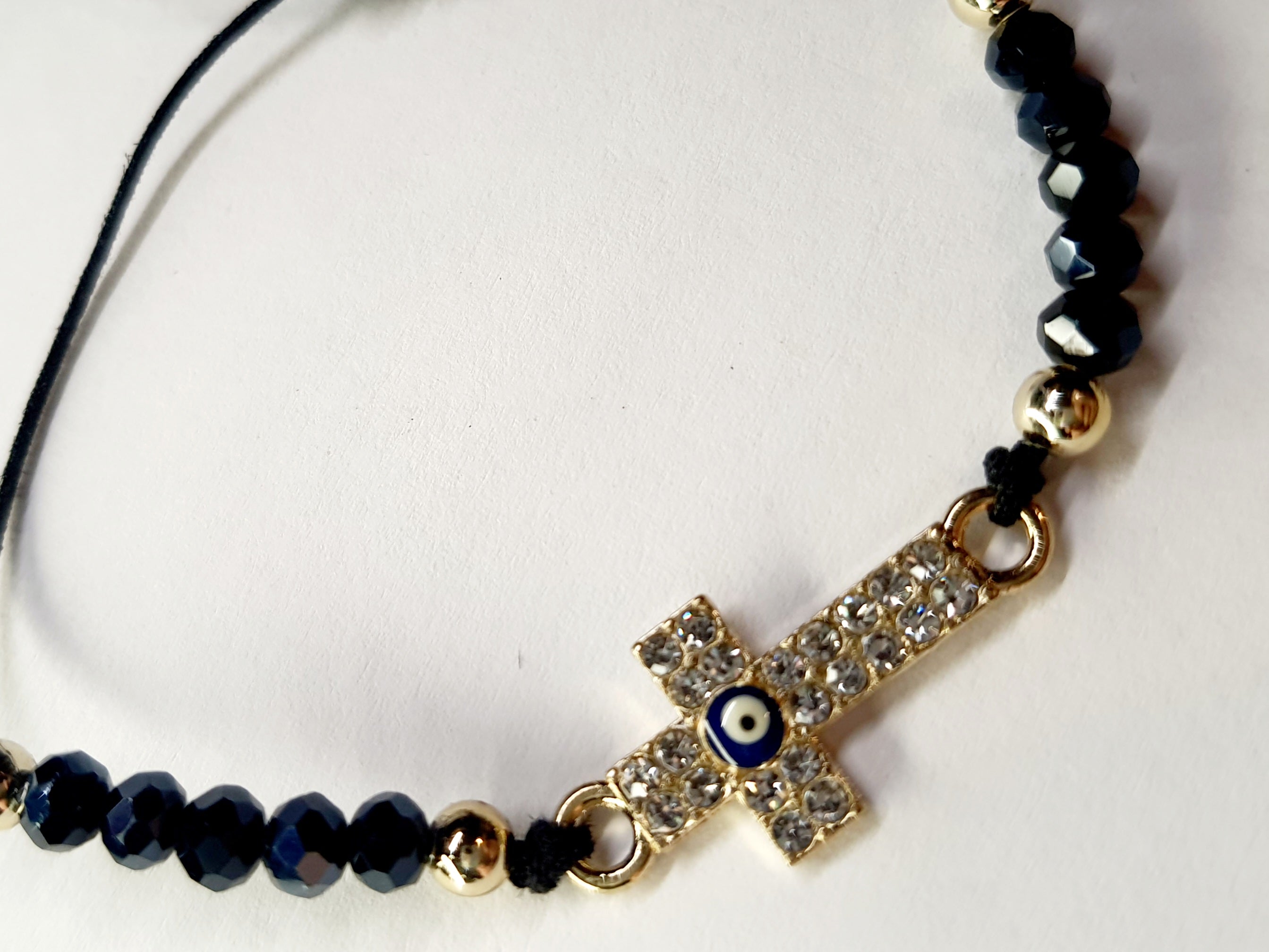 Bransoletka z krzyżem wysadzanym cyrkoniami i okiem proroka w złotej oprawie, na czarnym sznurku z ciemnymi koralikami. Ochronna i elegancka biżuteria w stylu boho. Wodoodporne, hipoalergiczne, jakość premium. Rouge Bizu.