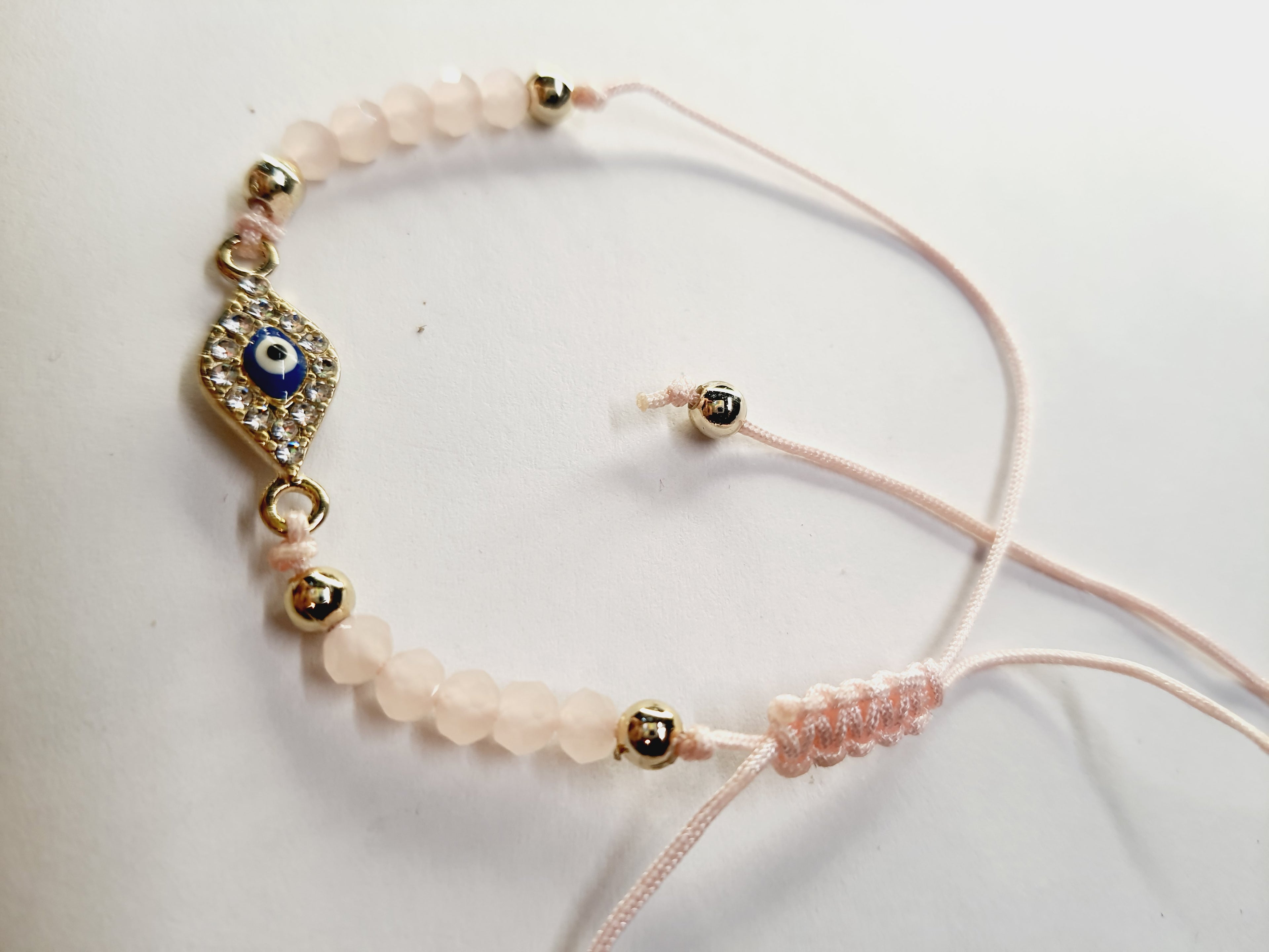 Bransoletka z okiem proroka w złotej oprawie wysadzanej cyrkoniami, na pudroworóżowym sznurku z matowymi różowymi koralikami. Delikatna, kobieca biżuteria ochronna w stylu boho. Wodoodporne, hipoalergiczne, jakość premium. Rouge Bizu.