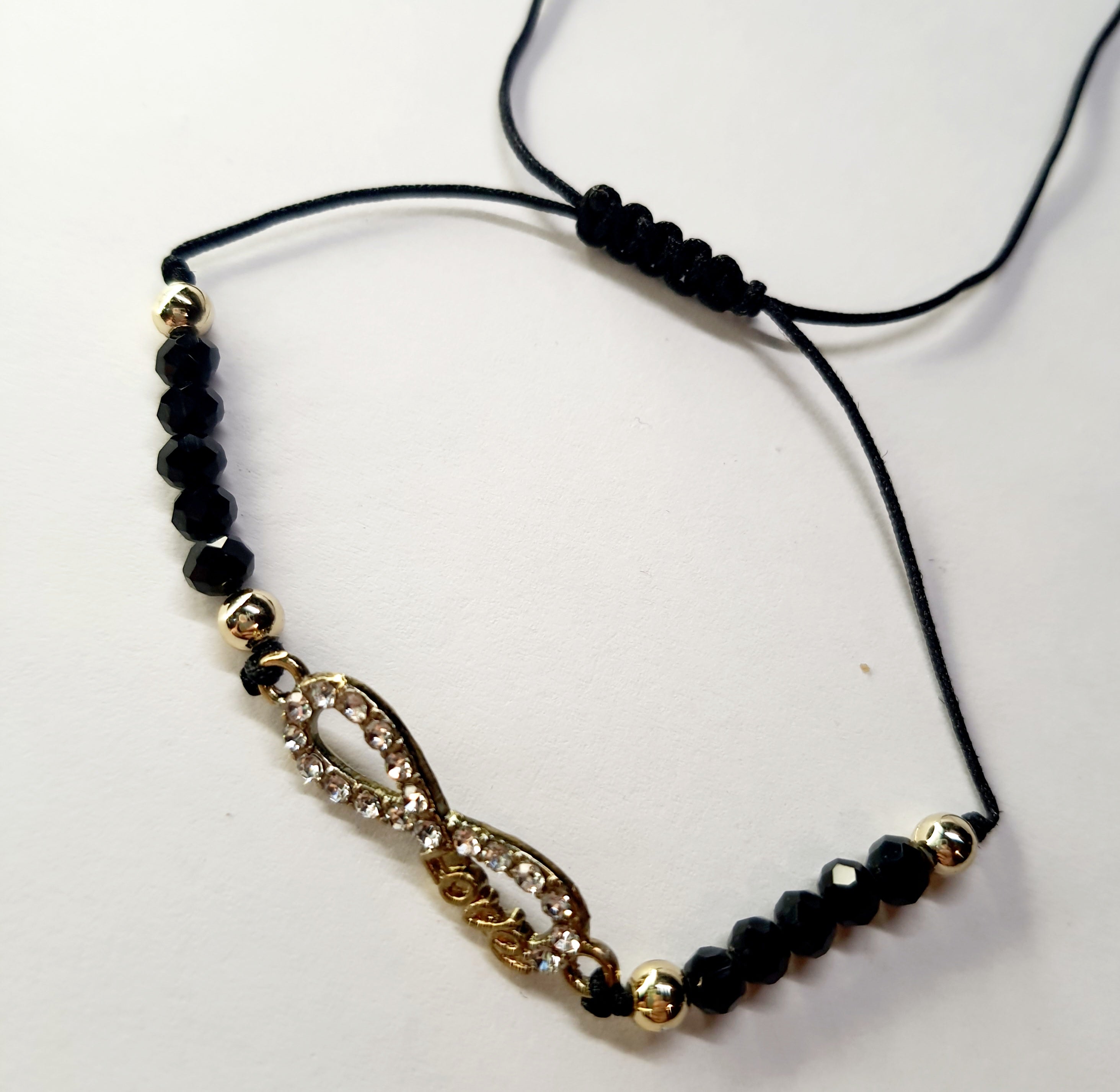 Bransoletka z symbolem nieskończoności z napisem „love”, wysadzana cyrkoniami, na czarnym sznurku z czarnymi koralikami. Elegancka, romantyczna biżuteria w złotym odcieniu. Wodoodporne, hipoalergiczne, jakość premium. Rouge Bizu.