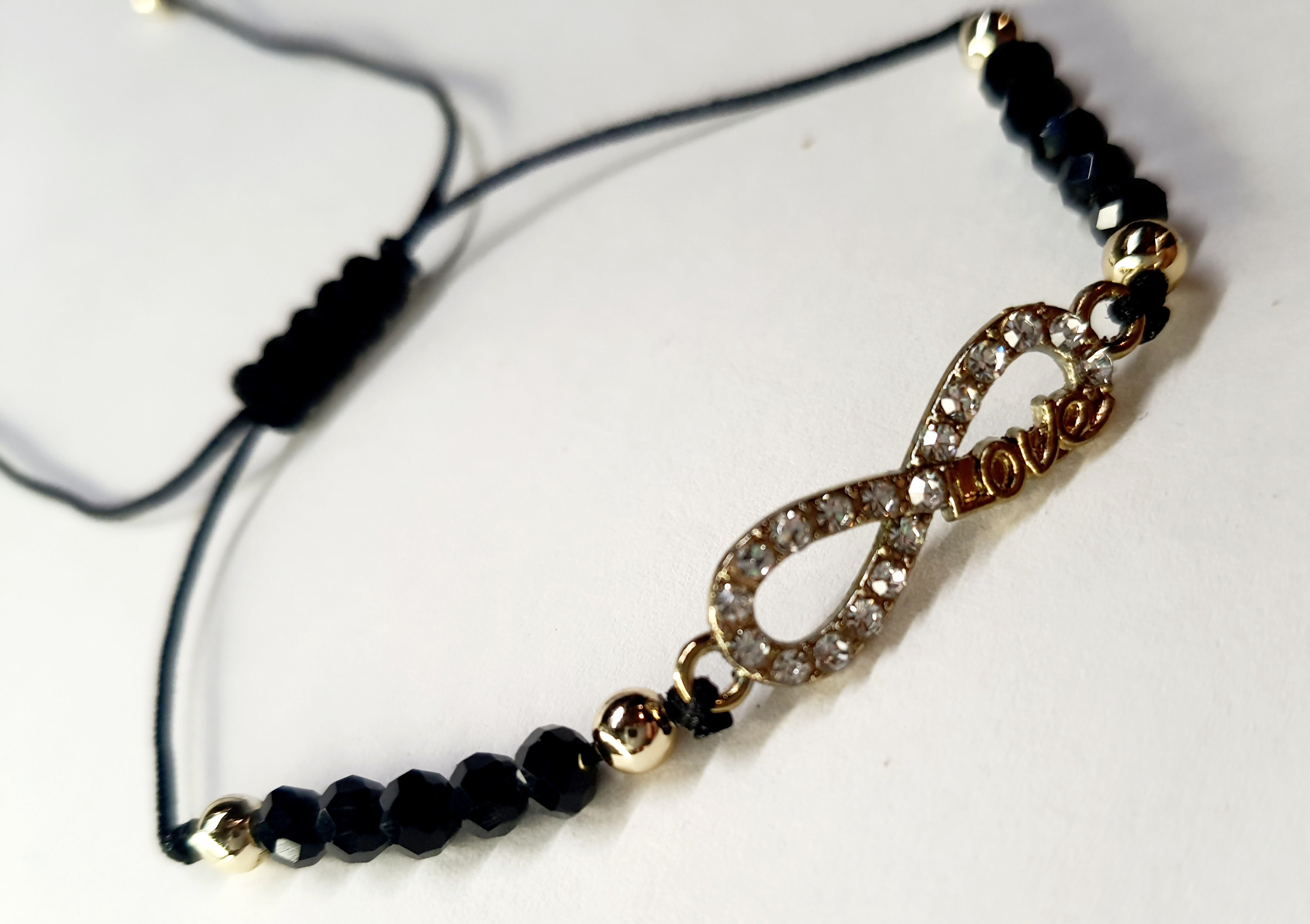 Bransoletka z symbolem nieskończoności z napisem „love”, wysadzana cyrkoniami, na czarnym sznurku z czarnymi koralikami. Elegancka, romantyczna biżuteria w złotym odcieniu. Wodoodporne, hipoalergiczne, jakość premium. Rouge Bizu.