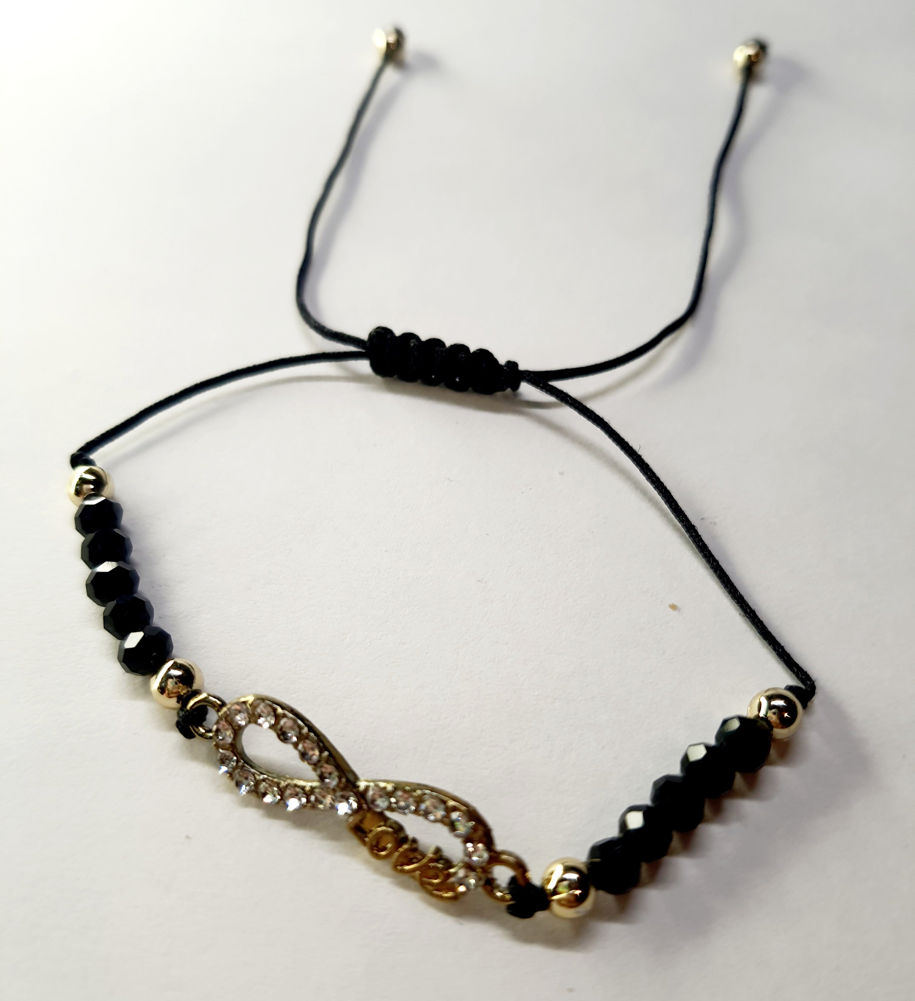 Bransoletka z symbolem nieskończoności z napisem „love”, wysadzana cyrkoniami, na czarnym sznurku z czarnymi koralikami. Elegancka, romantyczna biżuteria w złotym odcieniu. Wodoodporne, hipoalergiczne, jakość premium. Rouge Bizu.