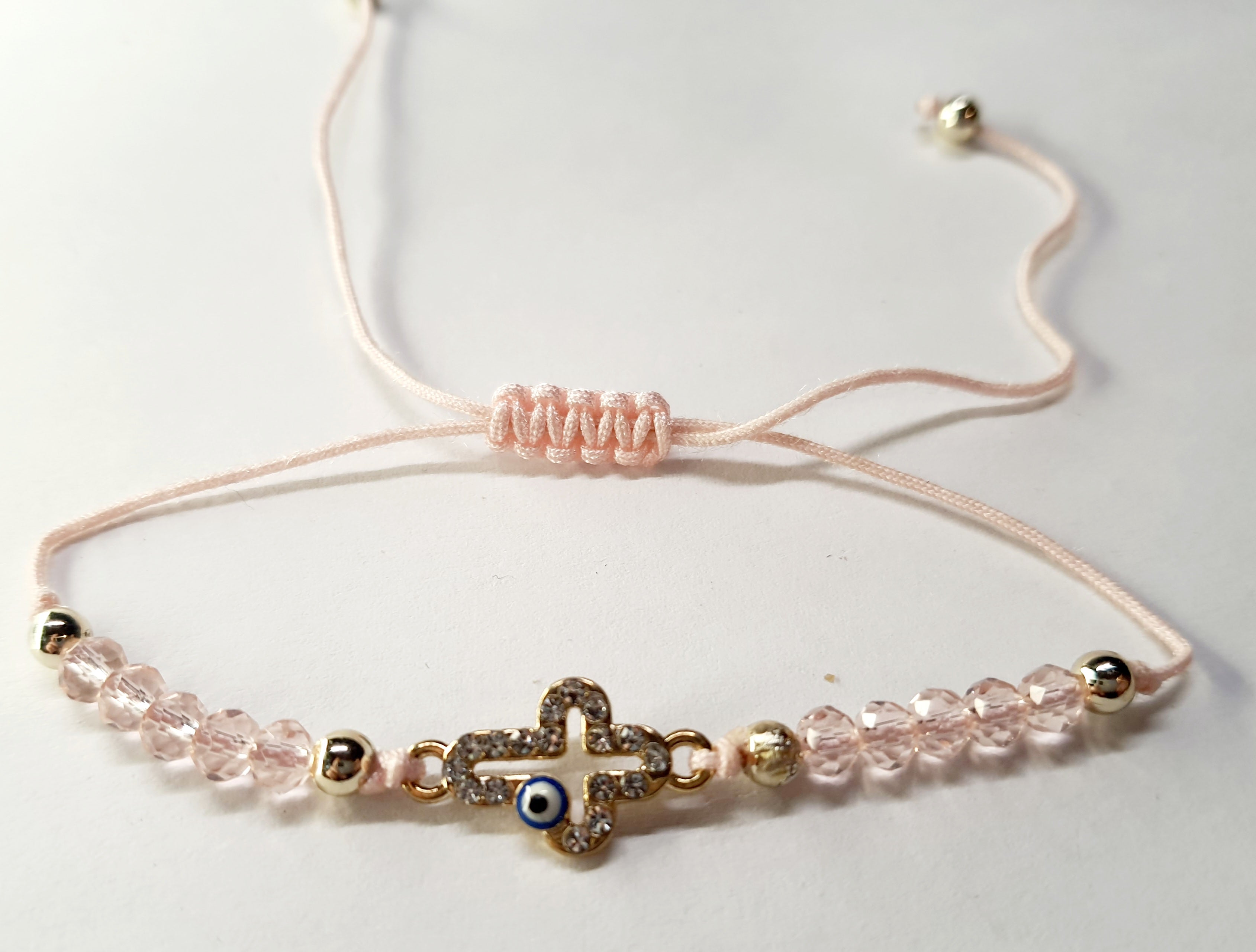 Bransoletka z krzyżem wysadzanym cyrkoniami i okiem proroka, na pudroworóżowym sznurku z różowymi koralikami. Delikatna, elegancka biżuteria ochronna w stylu boho. Wodoodporne, hipoalergiczne, jakość premium. Rouge Bizu.
