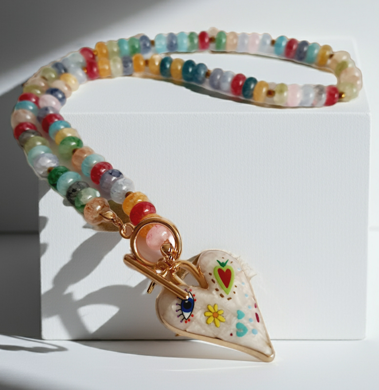 Naszyjnik z kolorowych koralików w stylu boho, z ozdobną zawieszką w kształcie białego serca zdobionego ręcznie malowanymi wzorami: oko, kwiat, serce i kolorowe symbole. Długość 45 cm. Stal nierdzewna. Rouge Bizu.