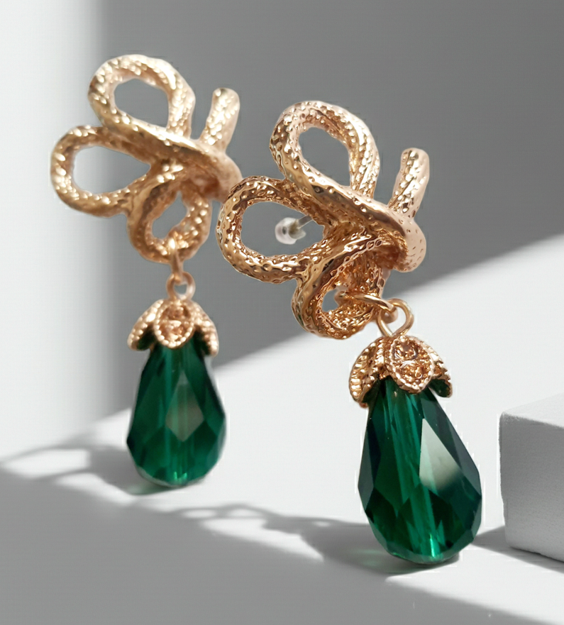 Złote kolczyki ze stali nierdzewnej w stylu glamour, ozdobione rzeźbioną kokardą i zielonym kryształem w kształcie kropli. Elegancja i szmaragdowy blask. Długość 6 cm. Rouge Bizu.