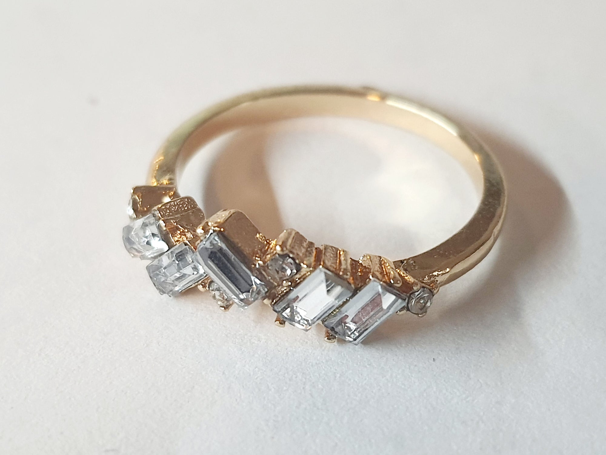 Pierścionek ze stali nierdzewnej w kolorze złotym, ozdobiony geometrycznymi przezroczystymi cyrkoniami w szlifie prostokątnym i kwadratowym. Styl glamour, rozmiar 16 mm. Rouge Bizu.