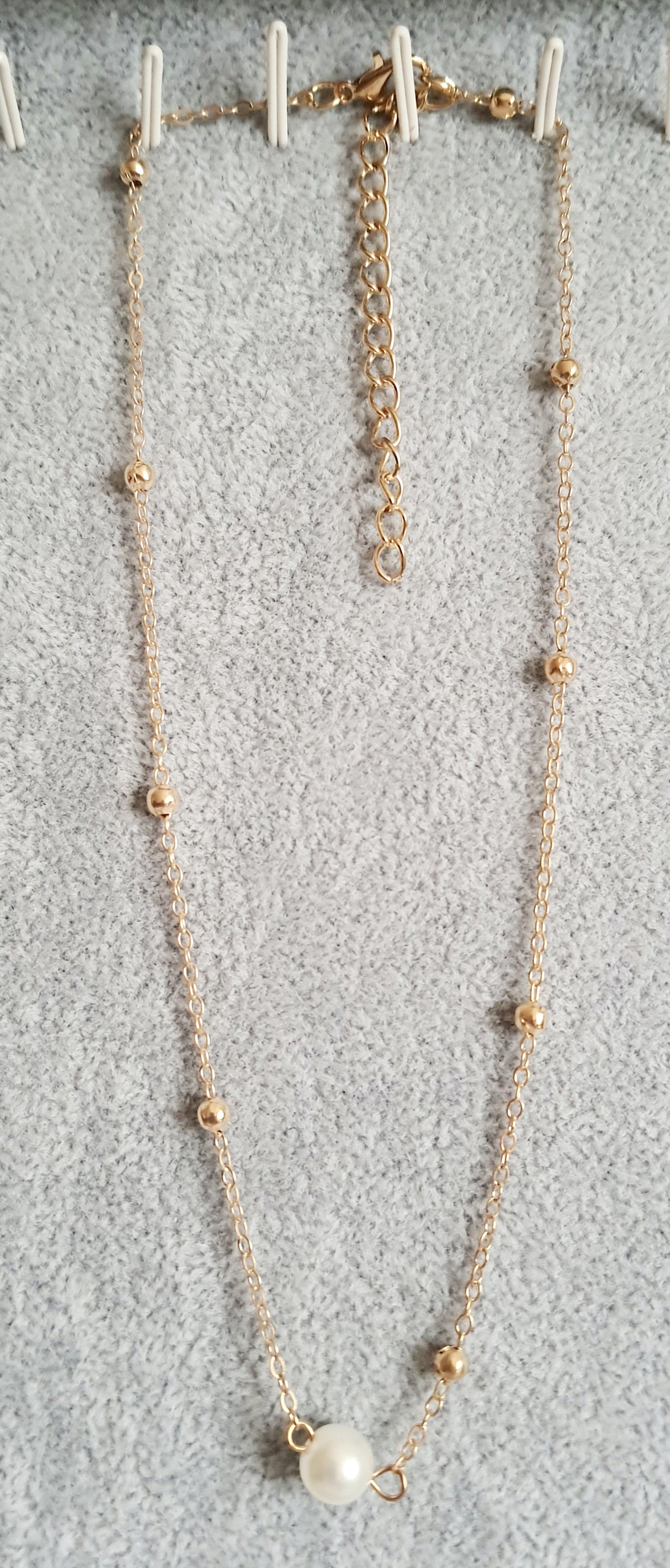 Złoty naszyjnik ze stali nierdzewnej z kulkami i zawieszką z białą perłą, ułożony na jasnym tle, typ celebrytka
ROUGEBIZU