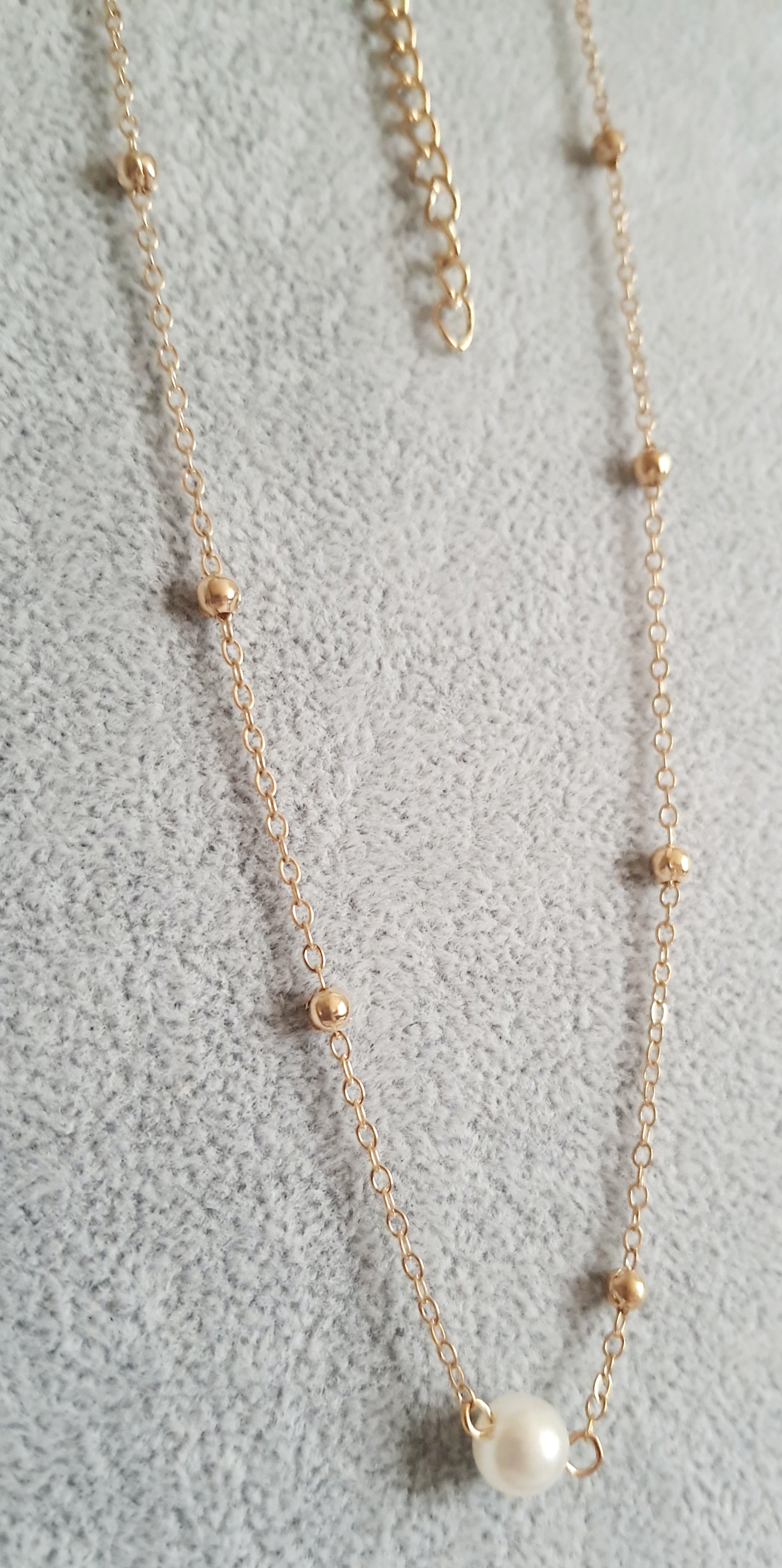 Złoty naszyjnik ze stali nierdzewnej z kulkami i zawieszką z białą perłą, ułożony na jasnym tle, typ celebrytka
ROUGEBIZU