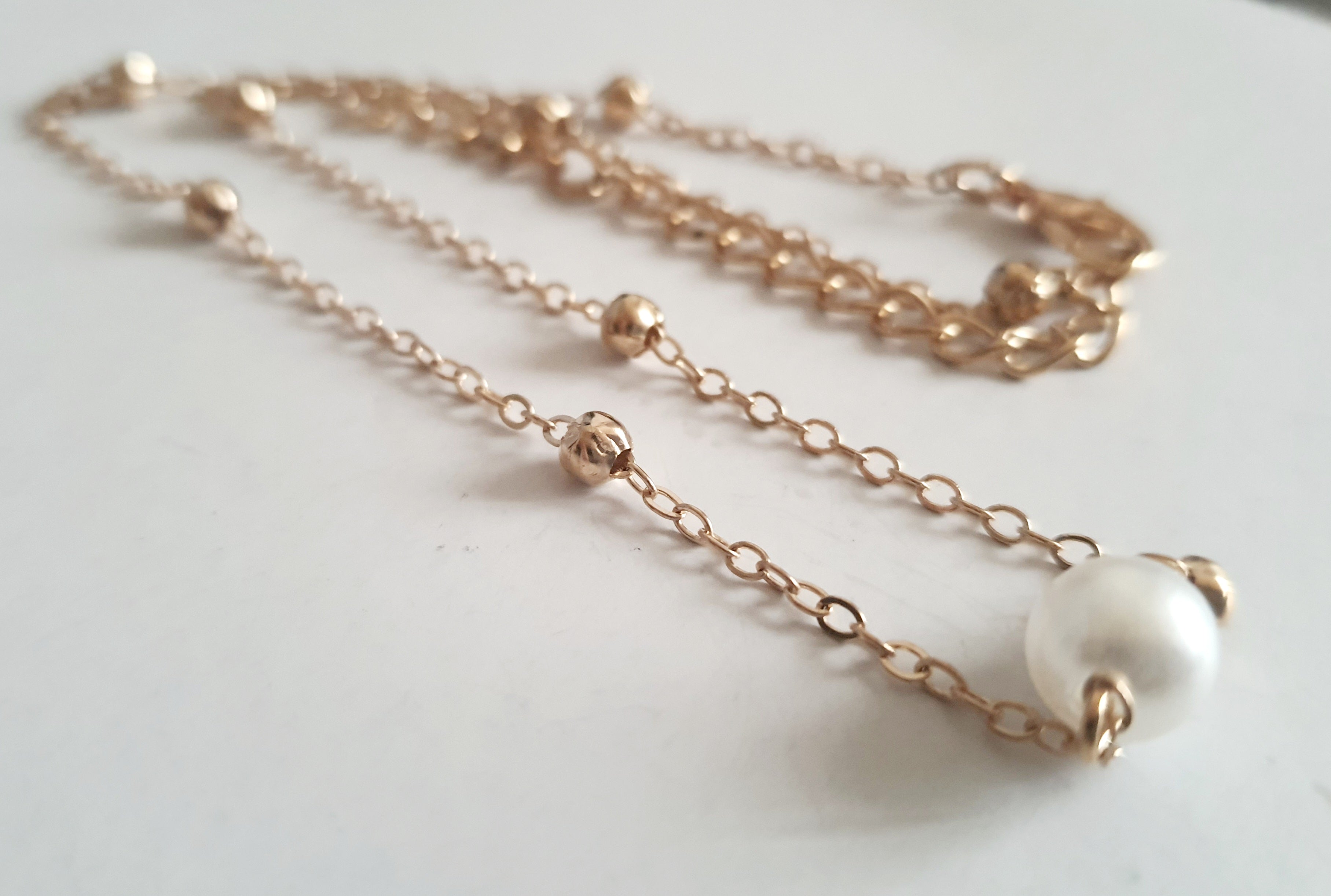 Złoty naszyjnik ze stali nierdzewnej z kulkami i zawieszką z białą perłą, ułożony na jasnym tle, typ celebrytka
ROUGEBIZU