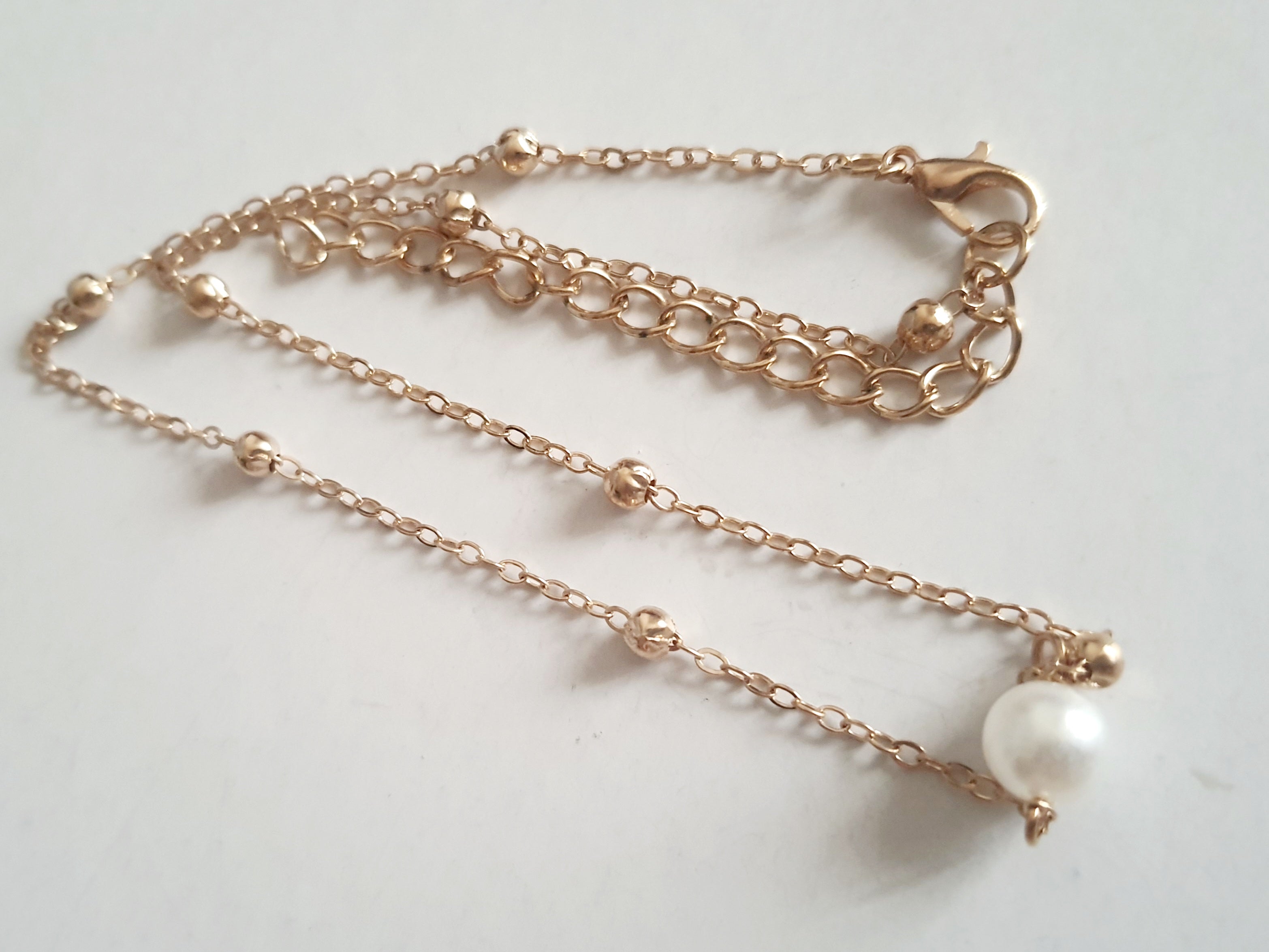 Złoty naszyjnik ze stali nierdzewnej z kulkami i zawieszką z białą perłą, ułożony na jasnym tle, typ celebrytka
ROUGEBIZU