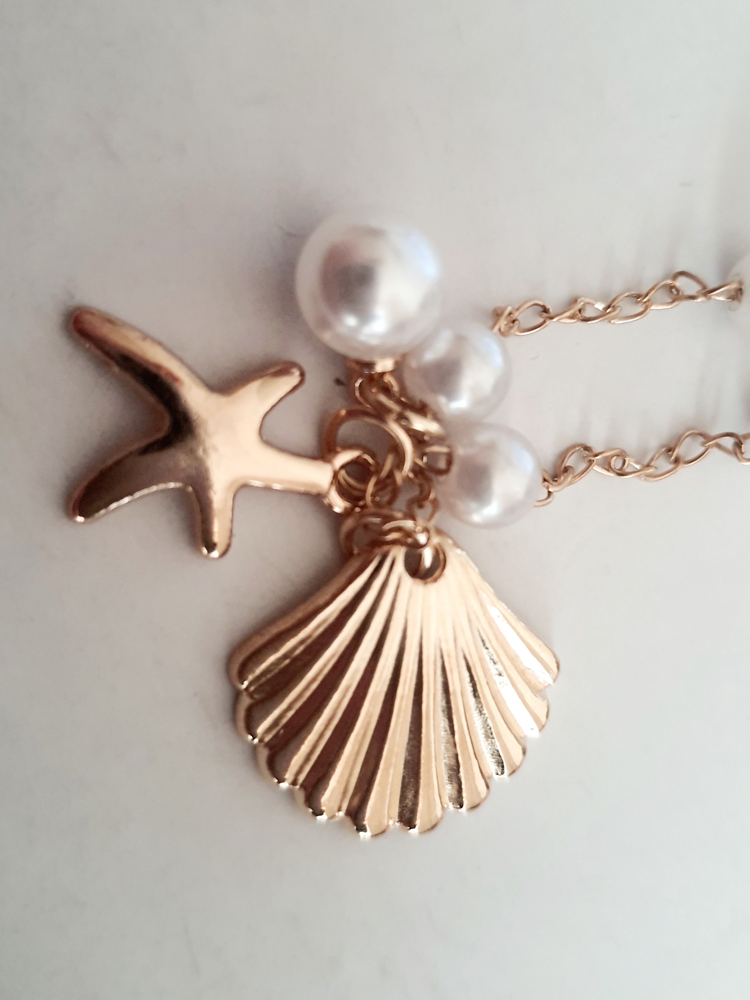 Naszyjnik z drobnymi syntetycznymi perełkami i złotymi zawieszkami w kształcie muszelki i rozgwiazdy. Długość regulowana 55–56 cm. Wykonany ze stali nierdzewnej (chirurgicznej). Delikatna biżuteria w stylu morskim. Rouge Bizu.

