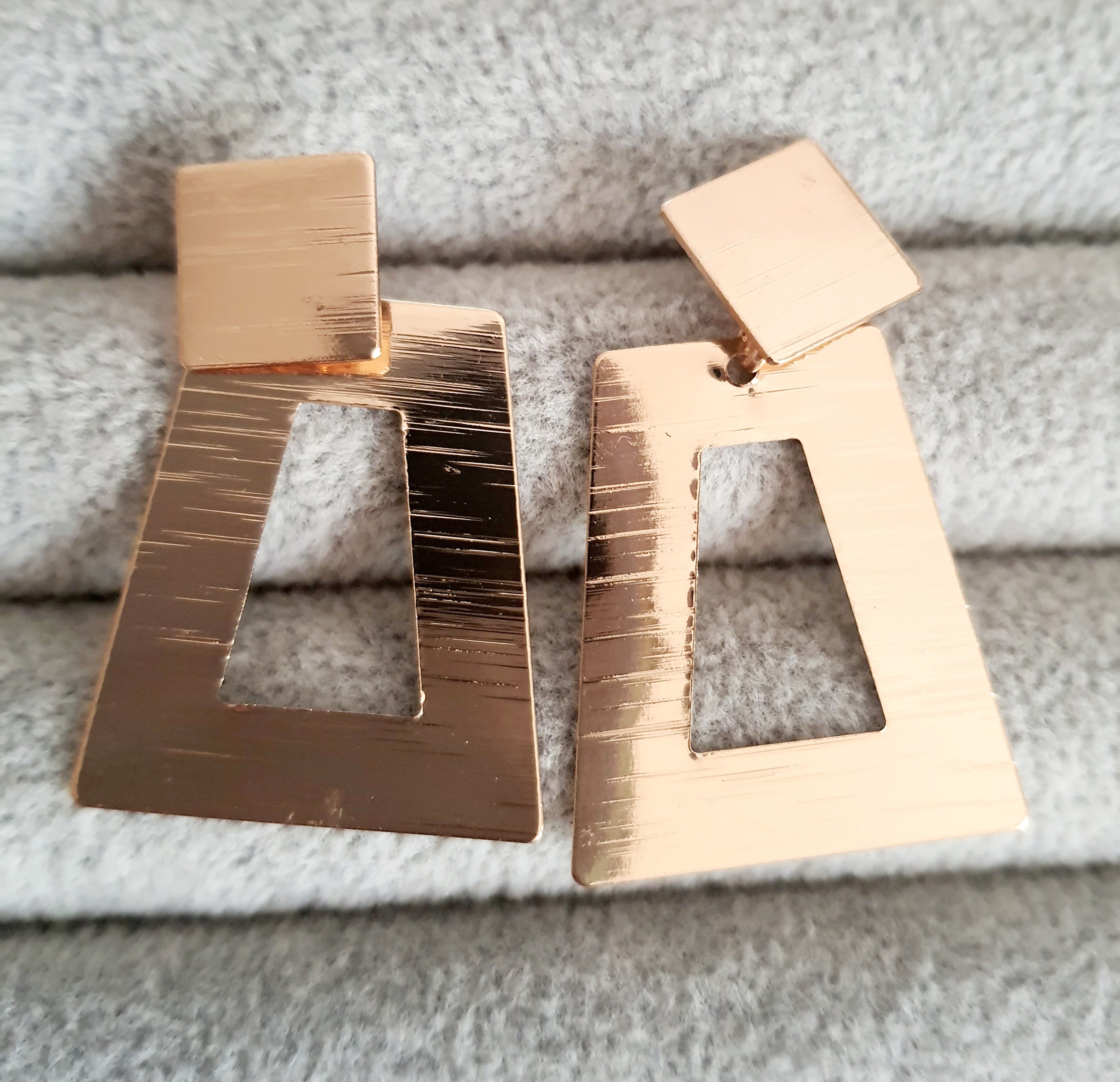 Geometryczne kolczyki z różowego złota o długości 5 cm, wykonane ze stali nierdzewnej. Trapez z wycięciem i kwadratowa baza w formie wkrętki. Nowoczesny, minimalistyczny design w stylu art deco. Rouge Bizu.

