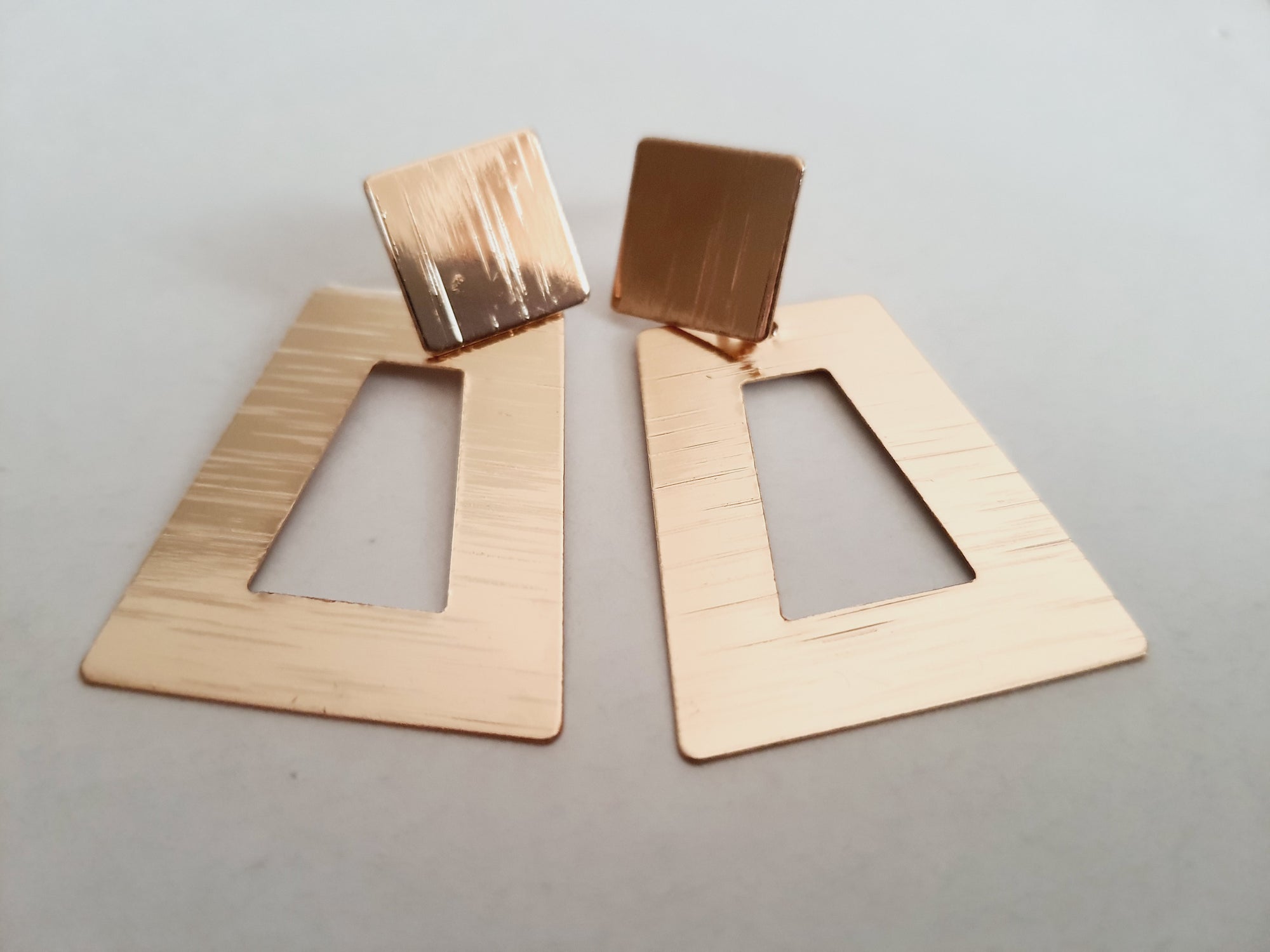 Geometryczne kolczyki z różowego złota o długości 5 cm, wykonane ze stali nierdzewnej. Trapez z wycięciem i kwadratowa baza w formie wkrętki. Nowoczesny, minimalistyczny design w stylu art deco. Rouge Bizu.

