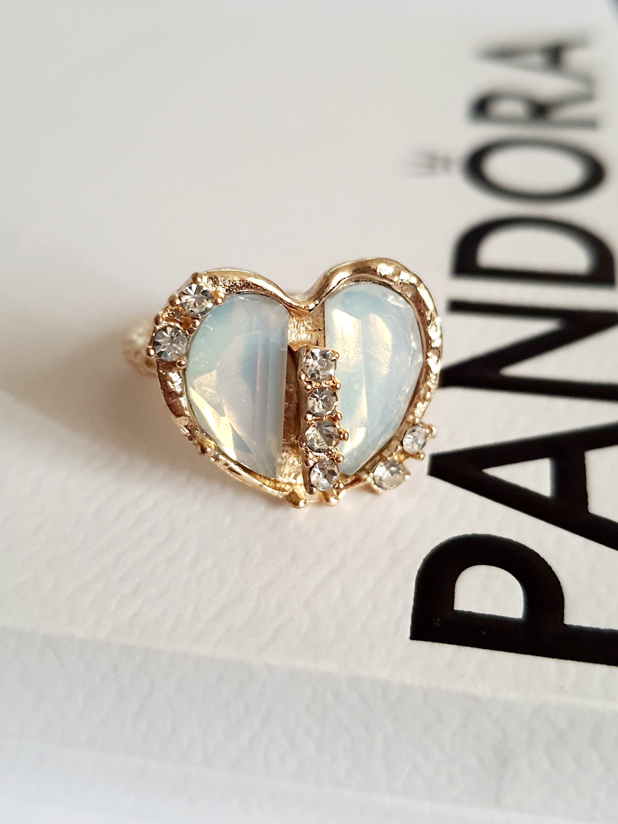 Romantyczny pierścionek ze stali nierdzewnej z podwójnym sercem z opalizującym, mlecznobiałym kamieniem i rzędem błyszczących cyrkonii. Styl glamour, rozmiar 17 mm, regulowany. Rouge Bizu.

