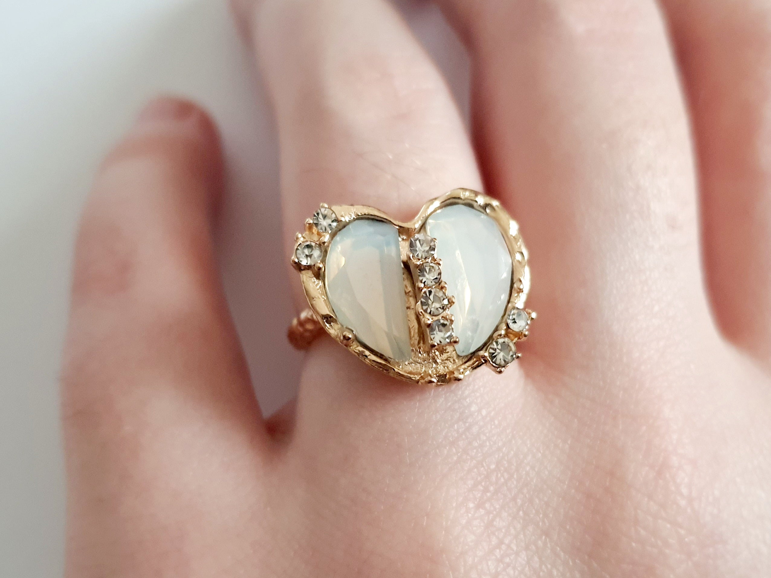 Romantyczny pierścionek ze stali nierdzewnej z podwójnym sercem z opalizującym, mlecznobiałym kamieniem i rzędem błyszczących cyrkonii. Styl glamour, rozmiar 17 mm, regulowany. Rouge Bizu.

