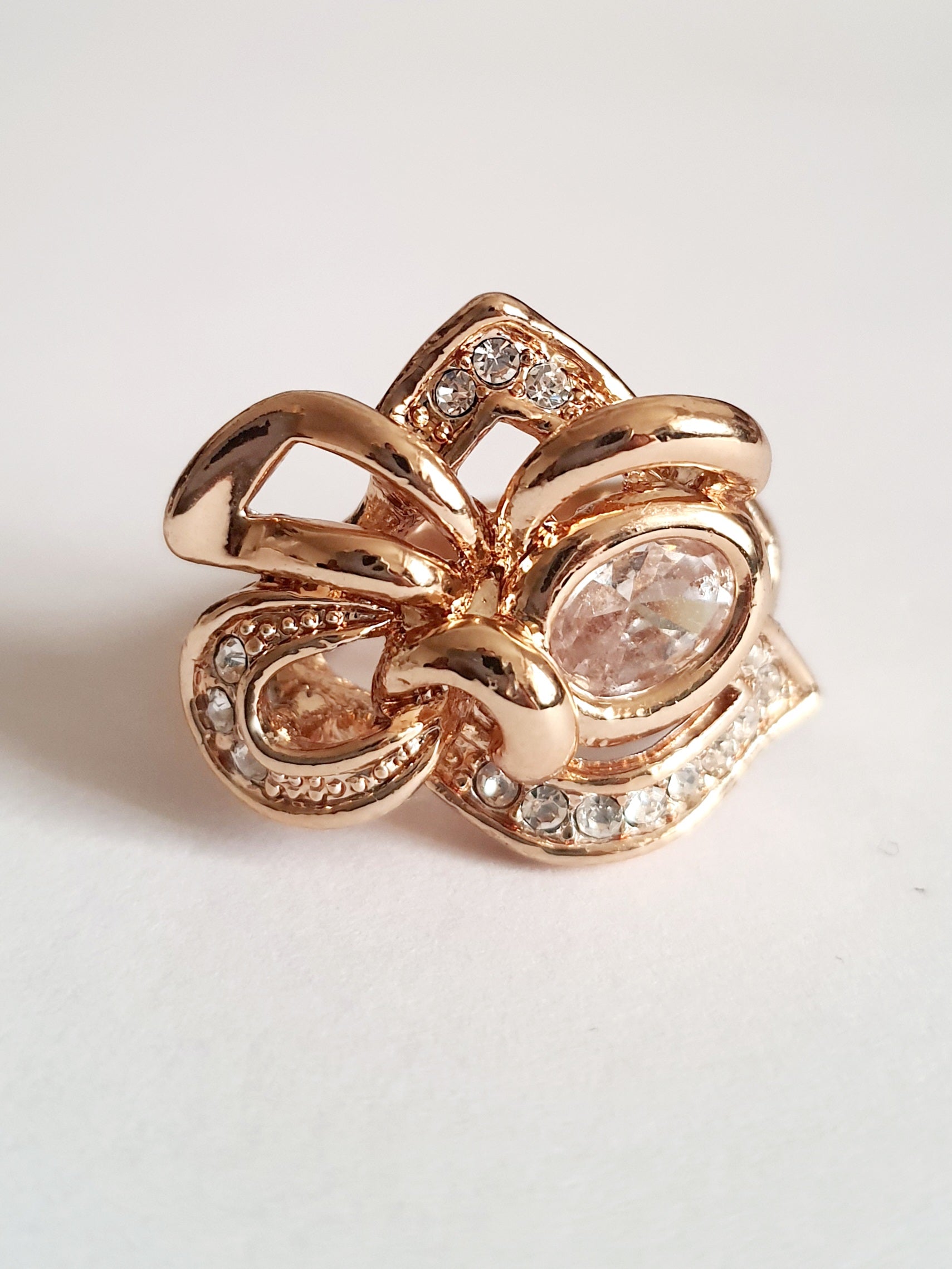 Pierścionek ze stali chirurgicznej w kolorze żółtego złota, z owalnym bezbarwnym kryształem i wijącymi się ramionami zdobionymi cyrkoniami. Styl glamour. Rozmiar 17 mm. Rouge Bizu.

