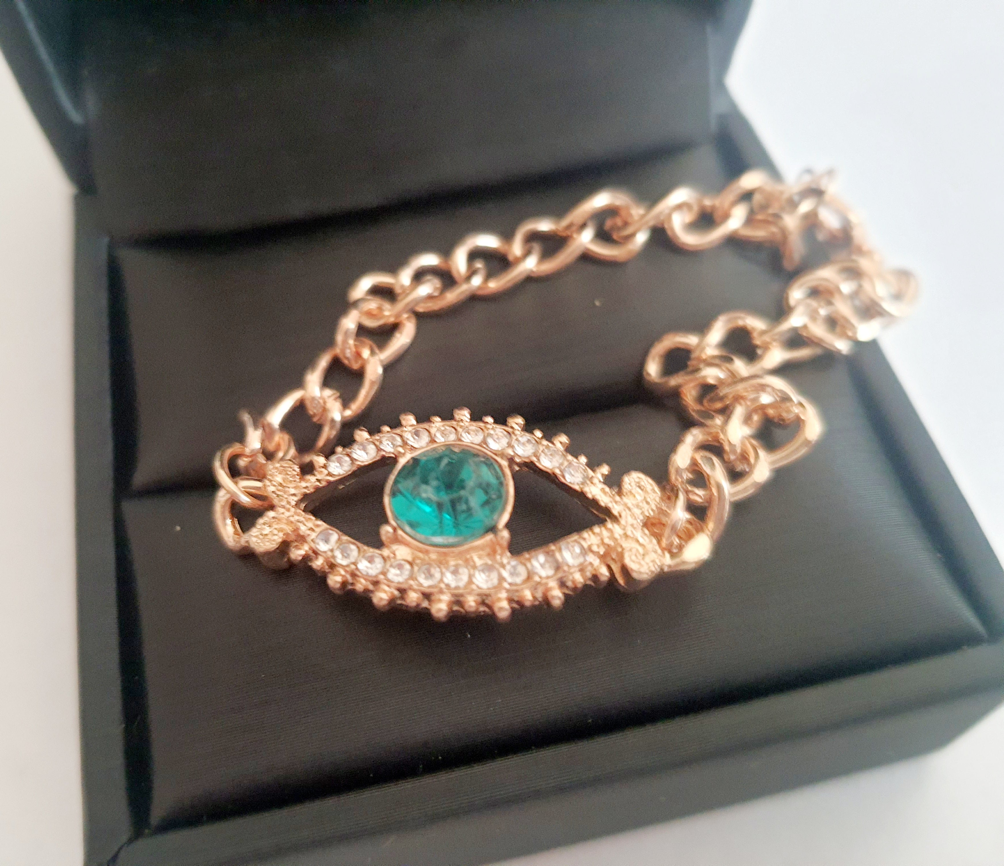 Bransoletka damska o długości 25–30 cm, złota, wykonana ze stali nierdzewnej. Ozdobiona motywem ochronnego oka z zieloną cyrkonią w centrum i białymi kamieniami wokół. Styl glamour z symbolicznym akcentem. Rouge Bizu.

