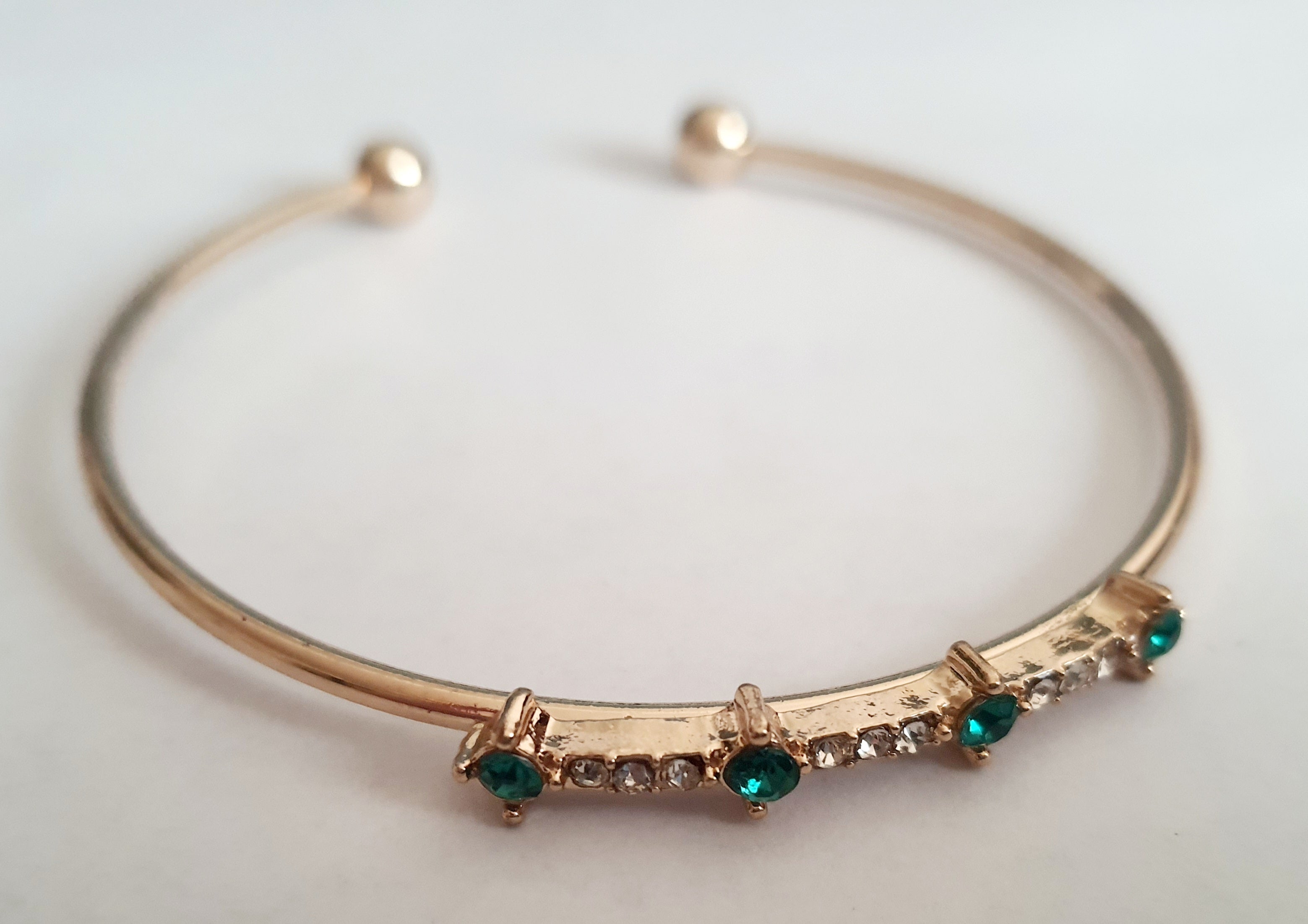 Sztywna złota bransoletka damska typu bangle ze stali nierdzewnej, ozdobiona szmaragdowymi kamieniami i drobnymi cyrkoniami. Styl glamour, elegancja i wyrazistość. Rouge Bizu.

