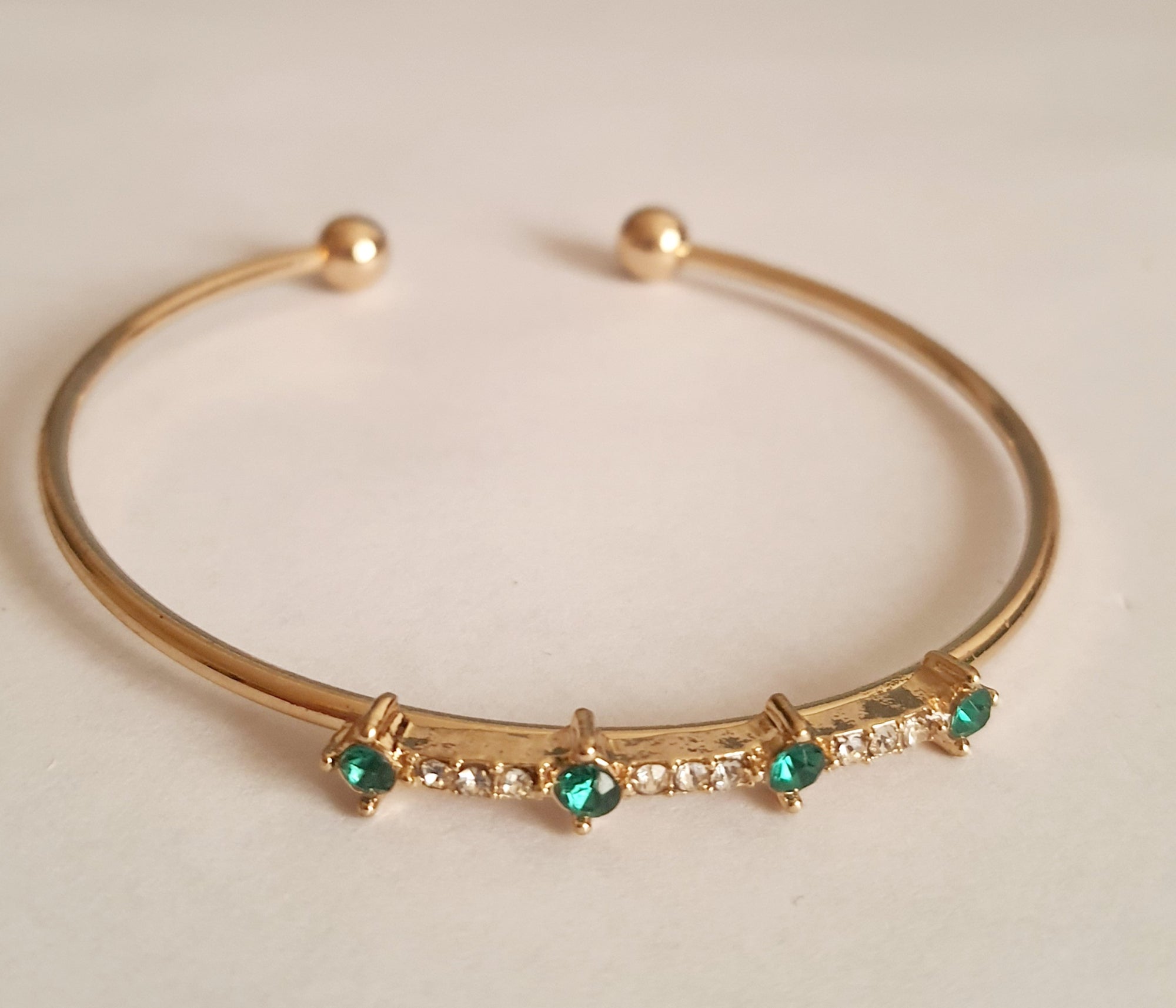 Sztywna złota bransoletka damska typu bangle ze stali nierdzewnej, ozdobiona szmaragdowymi kamieniami i drobnymi cyrkoniami. Styl glamour, elegancja i wyrazistość. Rouge Bizu.

