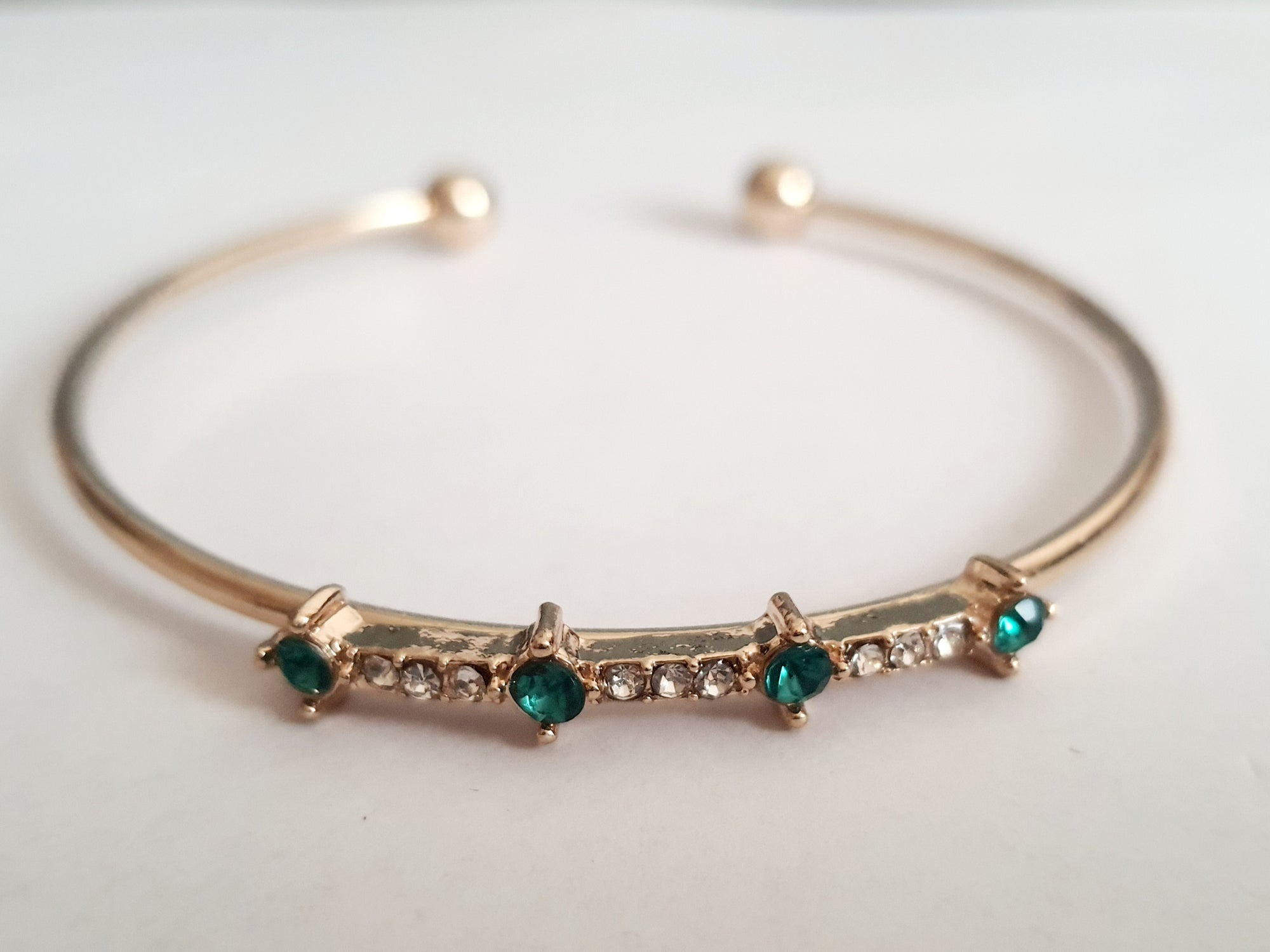 Sztywna złota bransoletka damska typu bangle ze stali nierdzewnej, ozdobiona szmaragdowymi kamieniami i drobnymi cyrkoniami. Styl glamour, elegancja i wyrazistość. Rouge Bizu.

