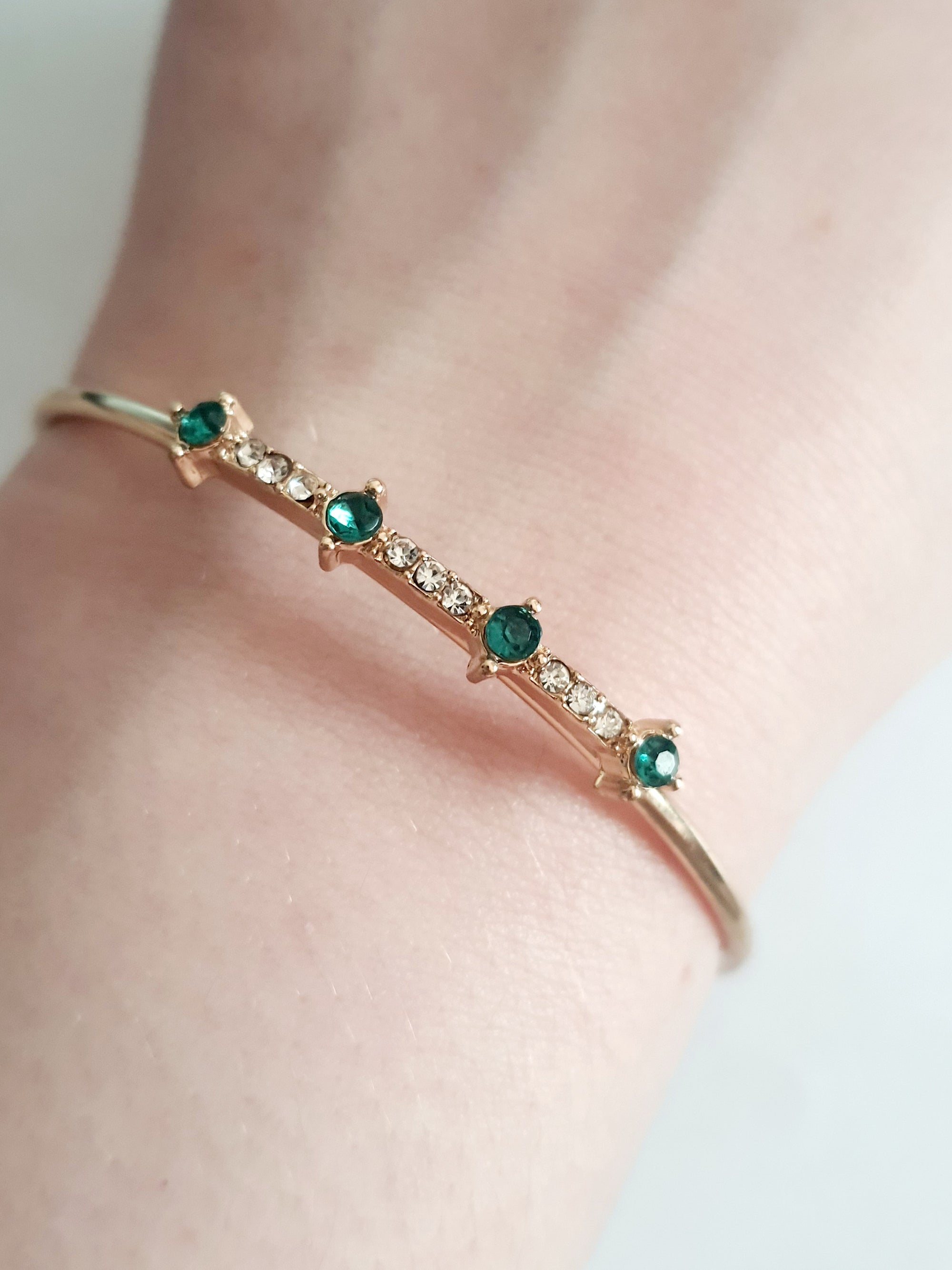 Sztywna złota bransoletka damska typu bangle ze stali nierdzewnej, ozdobiona szmaragdowymi kamieniami i drobnymi cyrkoniami. Styl glamour, elegancja i wyrazistość. Rouge Bizu.

