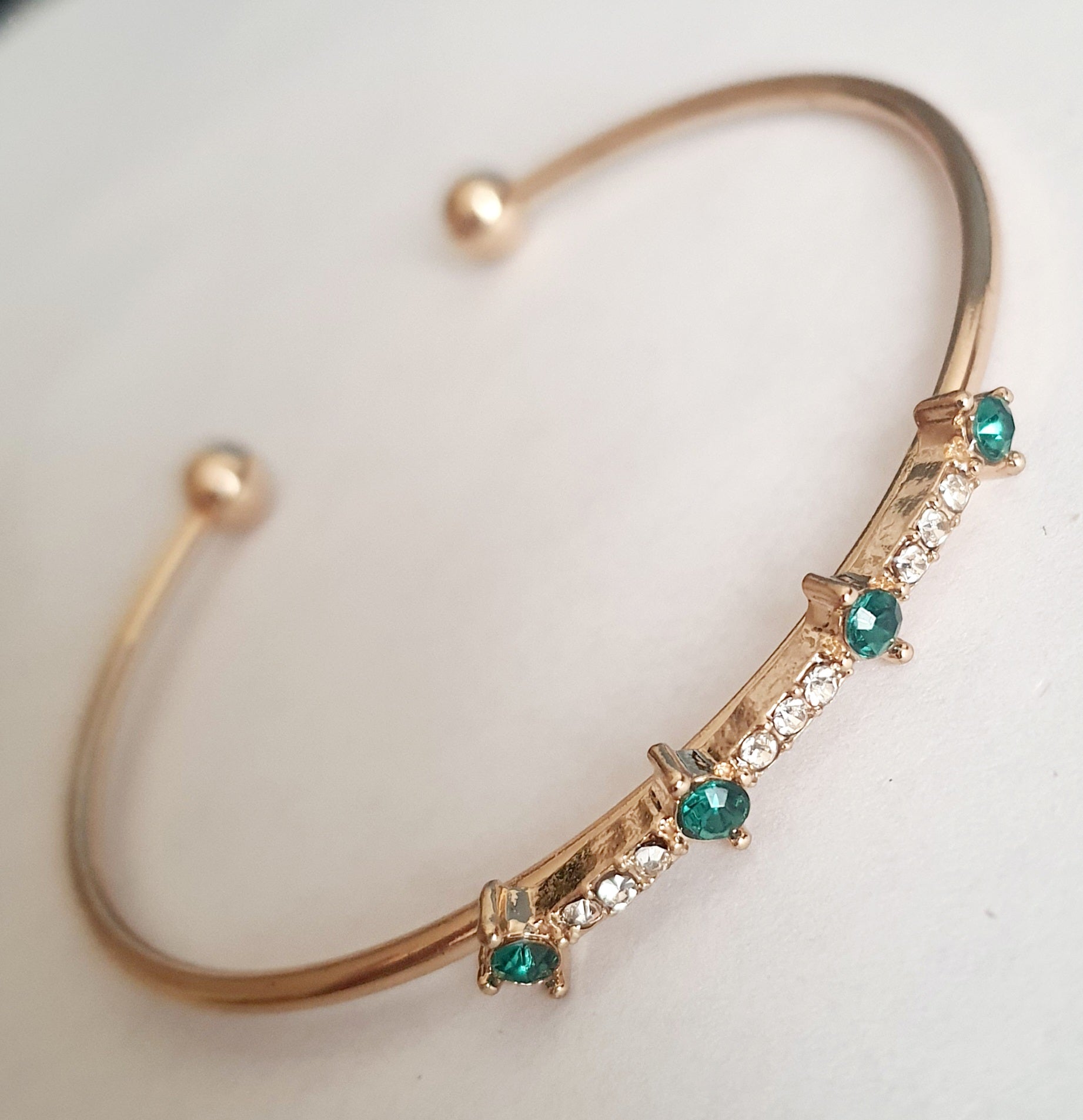 Sztywna złota bransoletka damska typu bangle ze stali nierdzewnej, ozdobiona szmaragdowymi kamieniami i drobnymi cyrkoniami. Styl glamour, elegancja i wyrazistość. Rouge Bizu.

