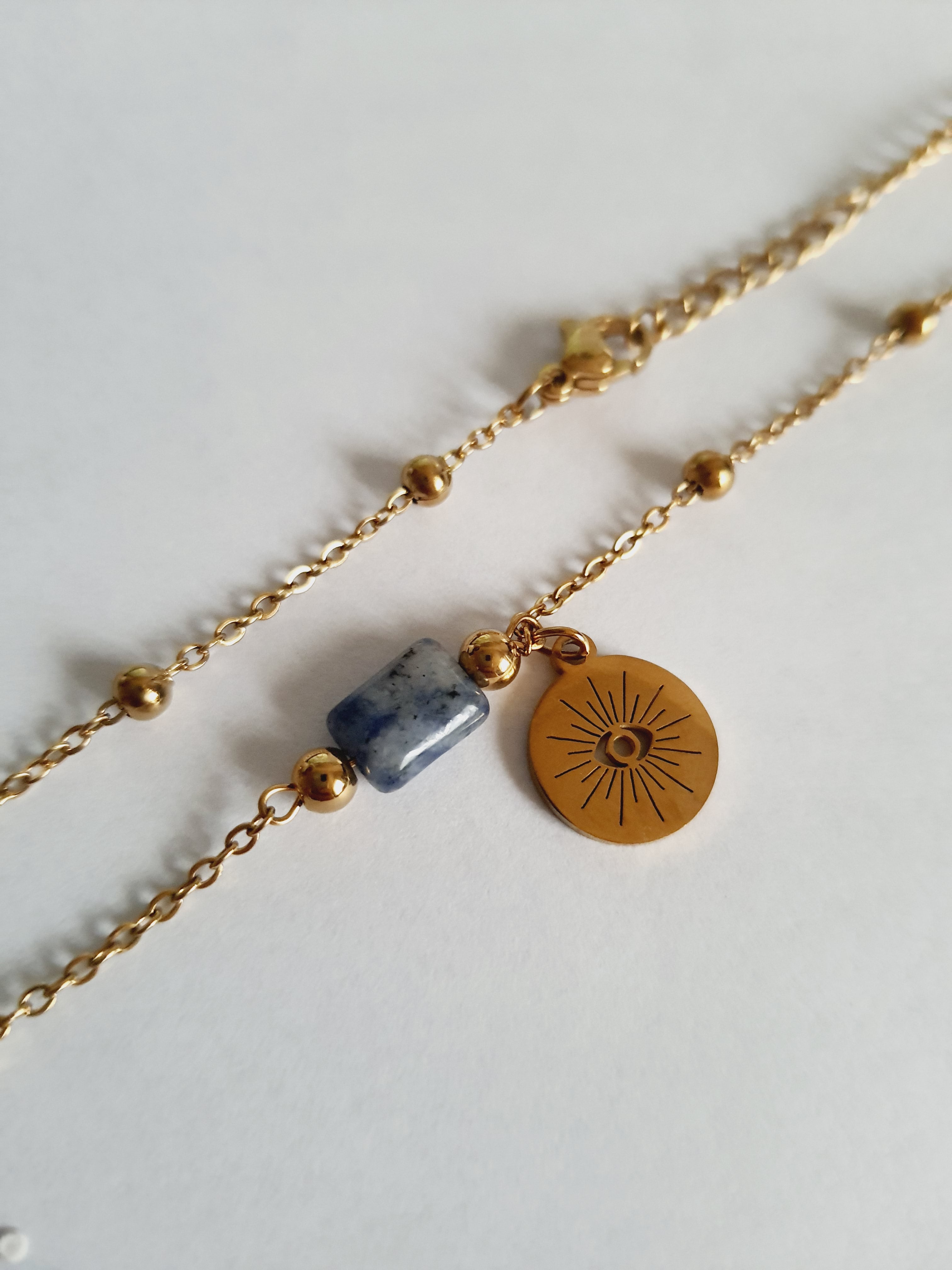 Złoty łańcuszek ze stali nierdzewnej o długości 40–45 cm z granatowym kamieniem i okrągłym talizmanem w kształcie słońca. Styl boho, minimalistyczny, duchowy. Rouge Bizu.

