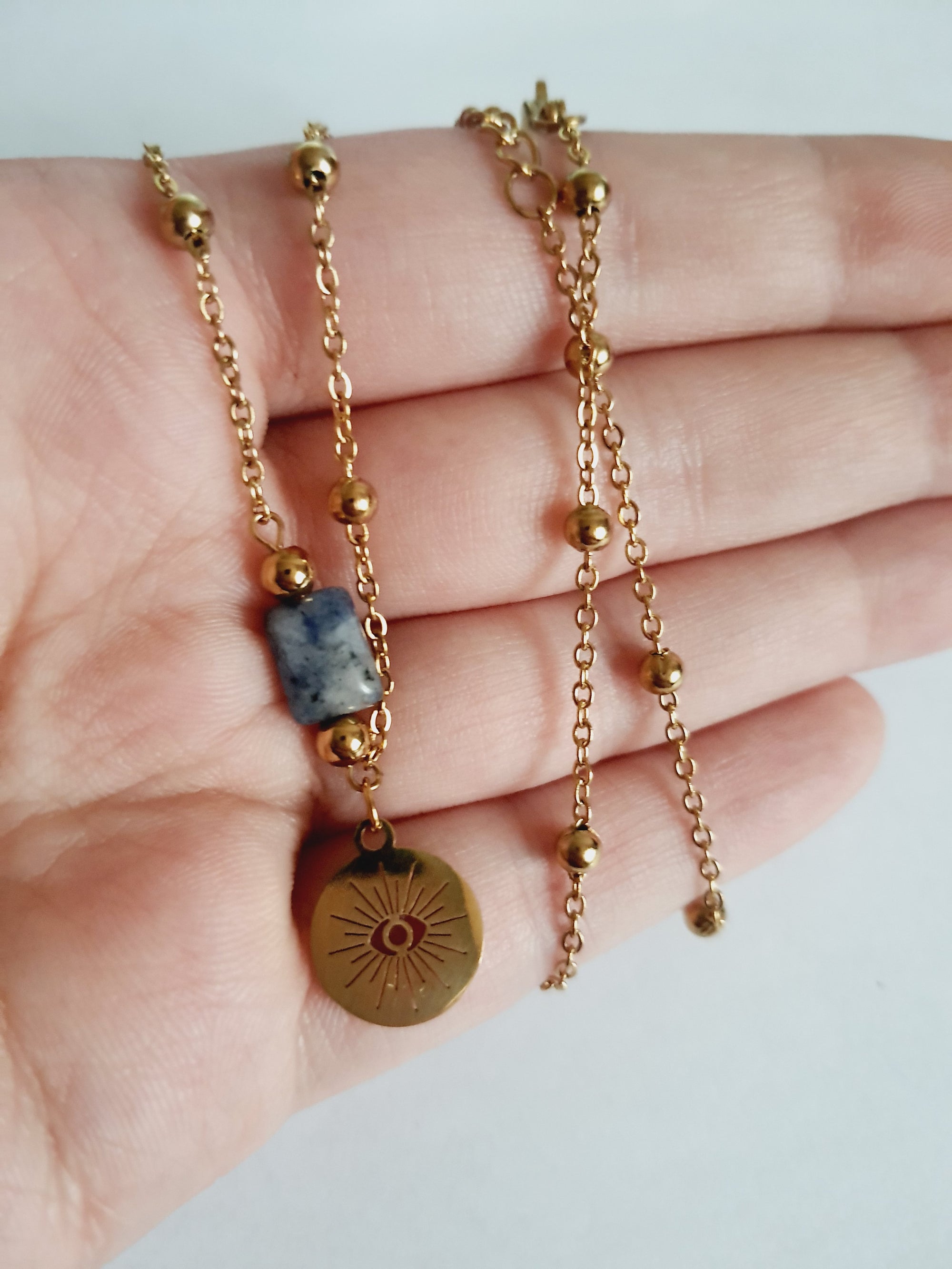 Złoty łańcuszek ze stali nierdzewnej o długości 40–45 cm z granatowym kamieniem i okrągłym talizmanem w kształcie słońca. Styl boho, minimalistyczny, duchowy. Rouge Bizu.

