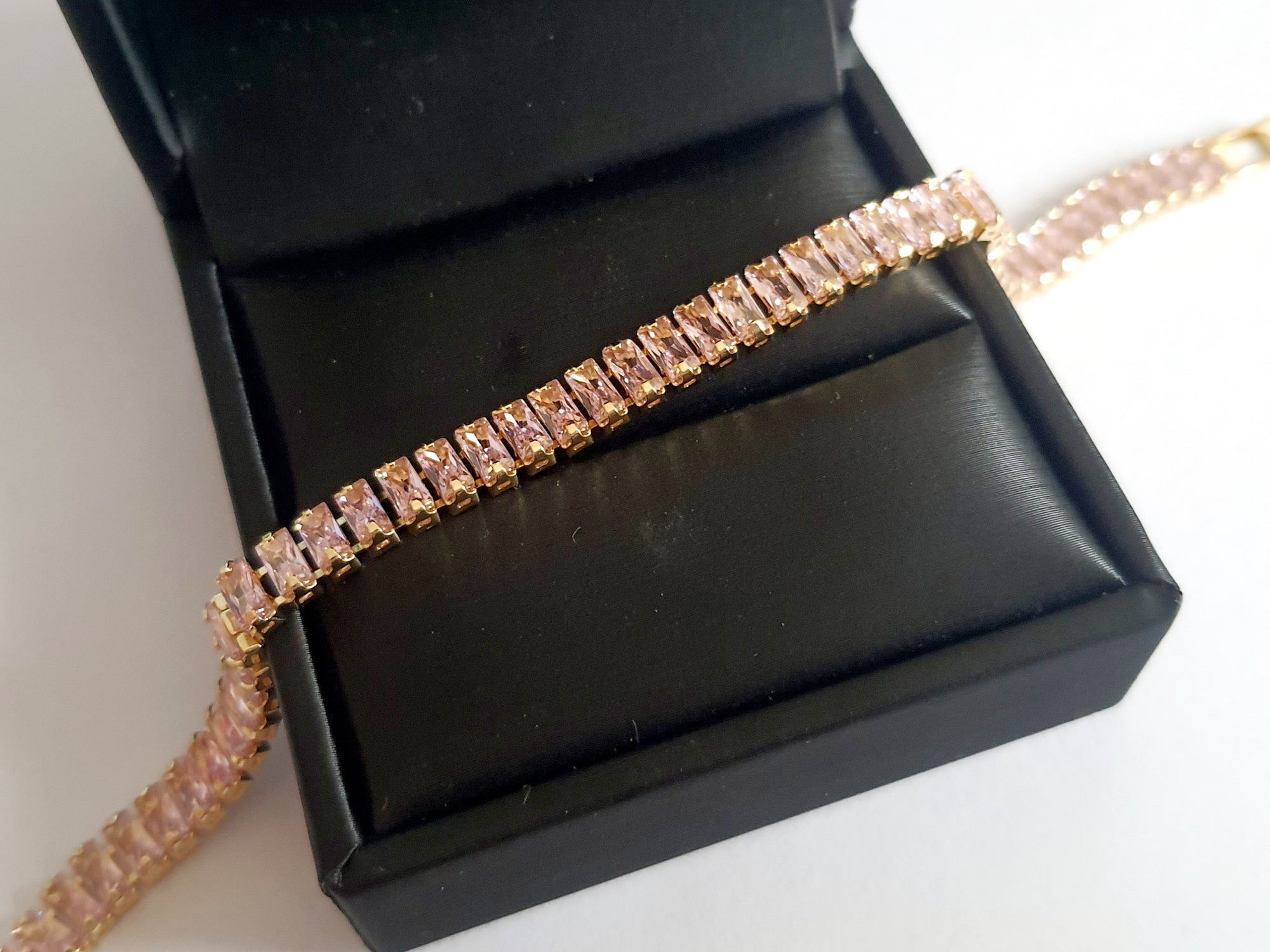 Bransoletka tenisowa ze stali nierdzewnej w złotej oprawie, zdobiona pudrowo-różowymi cyrkoniami, długość 19 cm. Klasyczna elegancja i romantyczny blask. Rouge Bizu.

