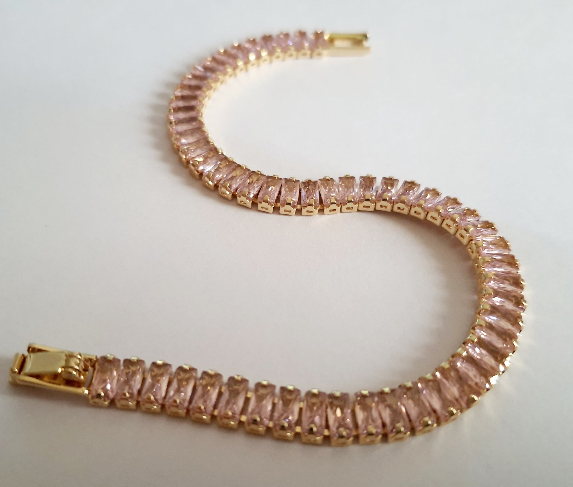 Bransoletka tenisowa ze stali nierdzewnej w złotej oprawie, zdobiona pudrowo-różowymi cyrkoniami, długość 19 cm. Klasyczna elegancja i romantyczny blask. Rouge Bizu.

