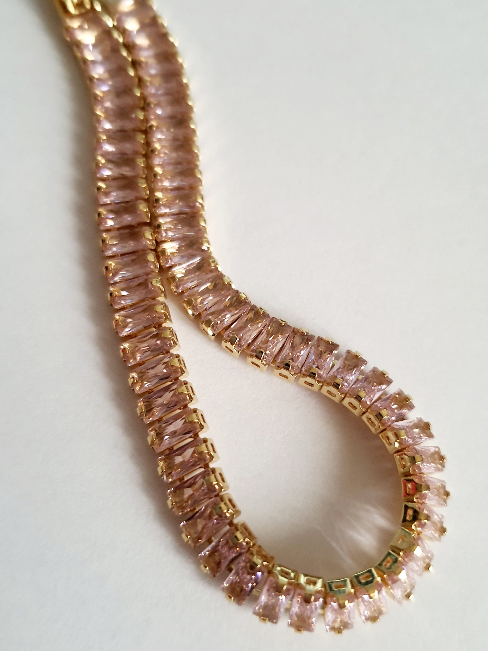 Bransoletka tenisowa ze stali nierdzewnej w złotej oprawie, zdobiona pudrowo-różowymi cyrkoniami, długość 19 cm. Klasyczna elegancja i romantyczny blask. Rouge Bizu.

